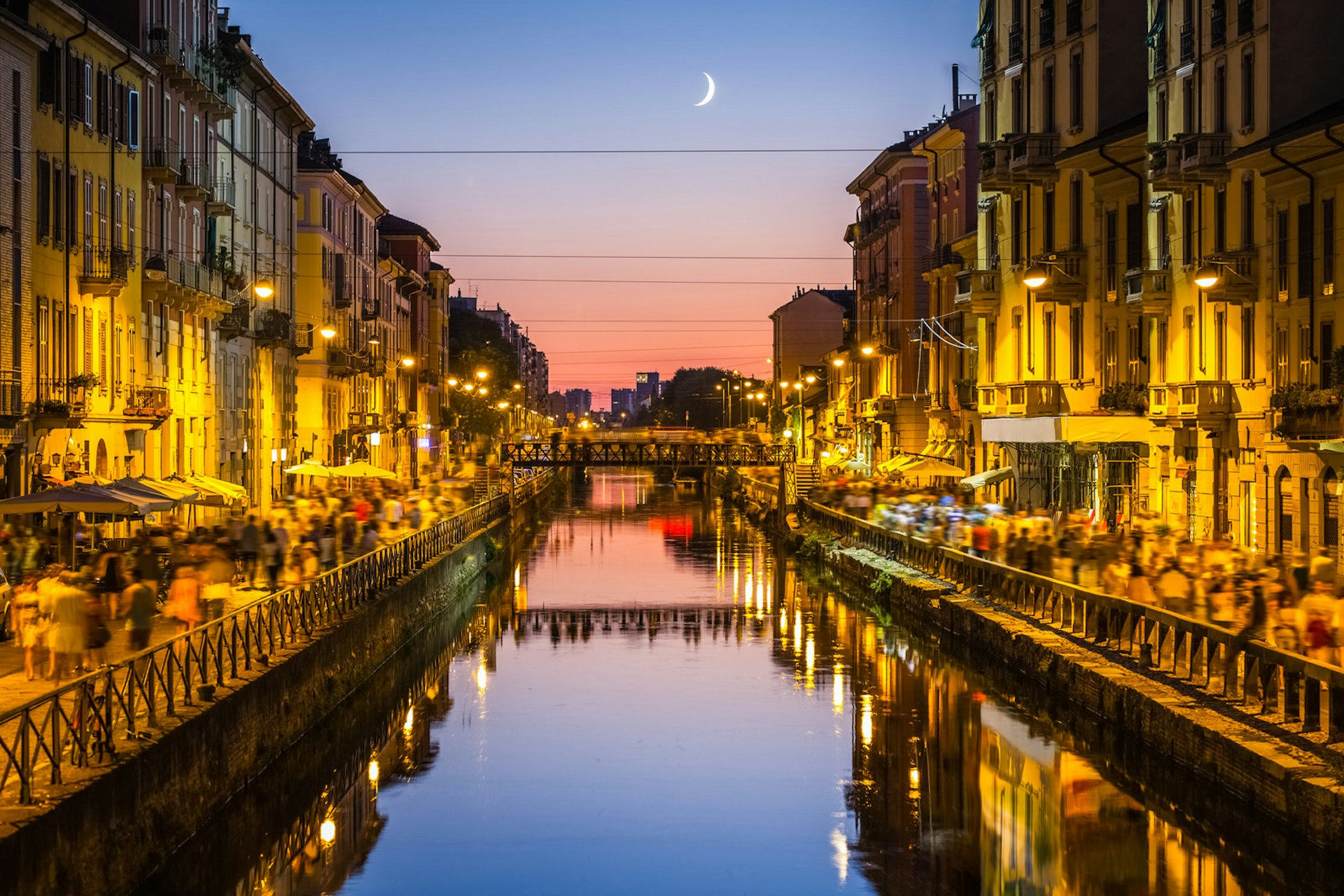 Sunset shot of a Navigli canal, with crowds of people walking down the streets on either side