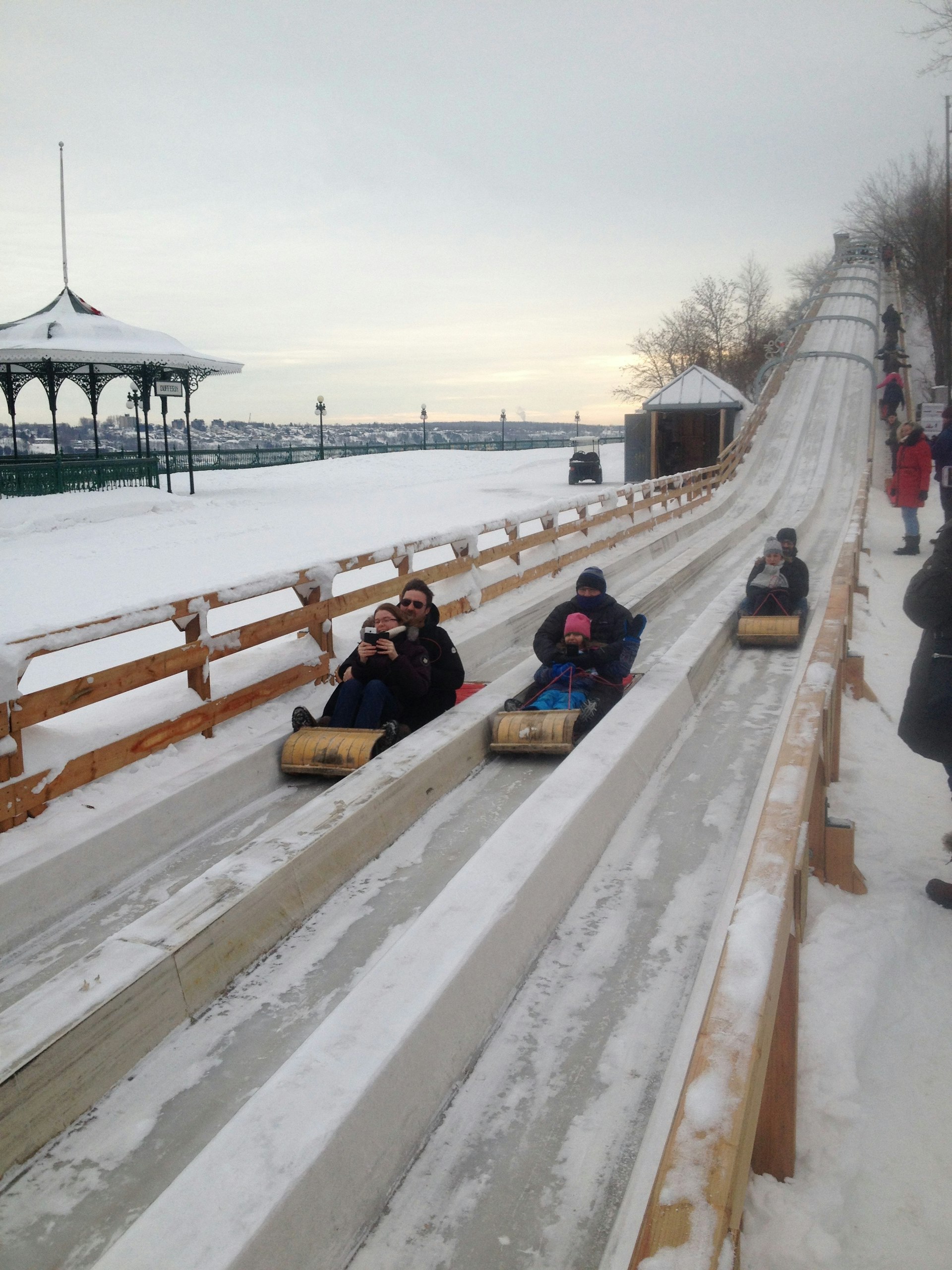 Three people sled down a large wooden slide toward the camera with the banks of a river to the left in Québec City.
