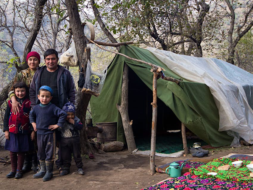 A Kyrgyz family poses in front of a makeshift green tent, with a picnic of bread on a blanket in the forest © Stephen Lioy / Lonely Planet
