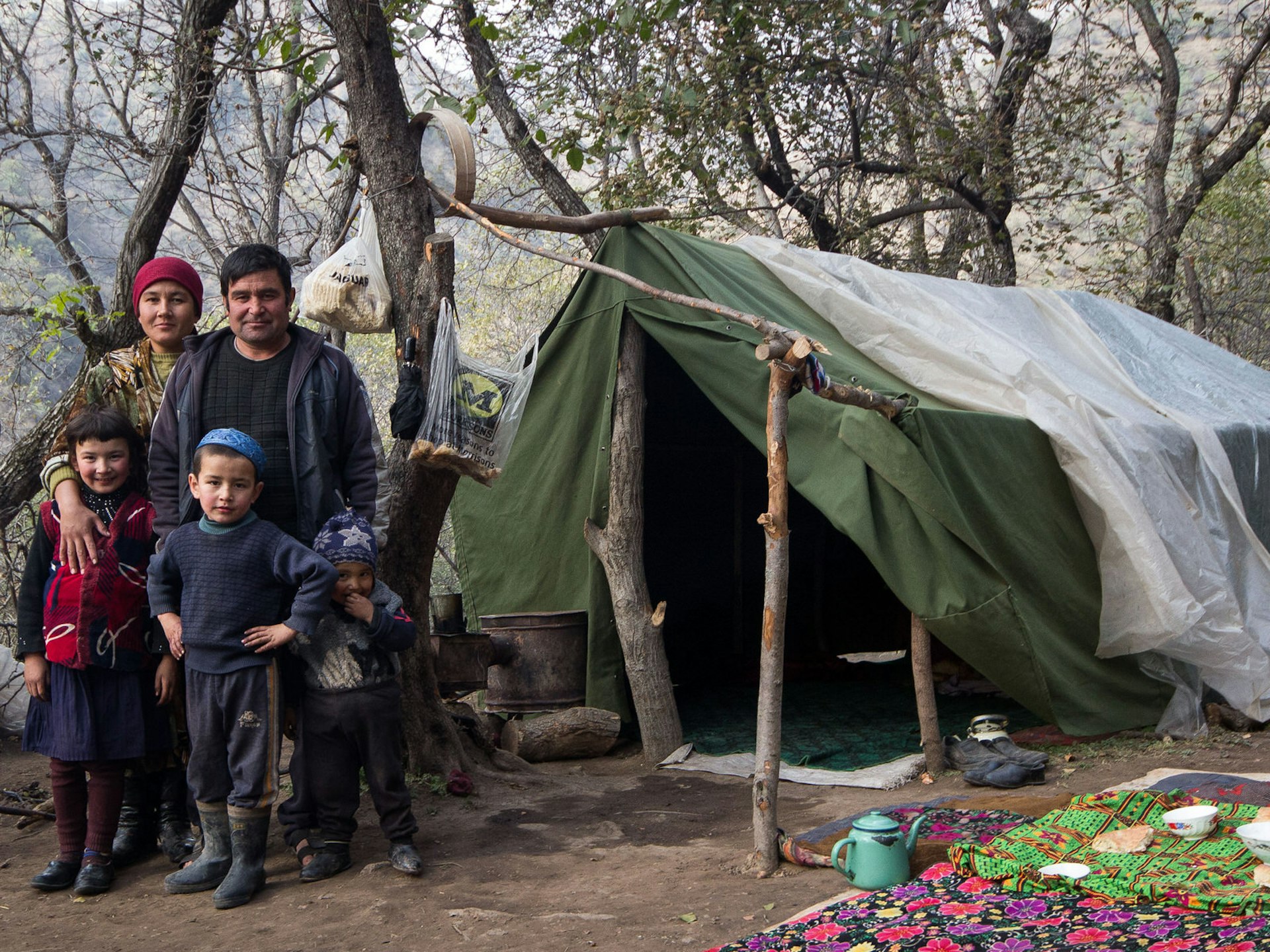 A Kyrgyz family poses in front of a makeshift green tent, with a picnic of bread on a blanket in the forest © Stephen Lioy / Lonely Planet