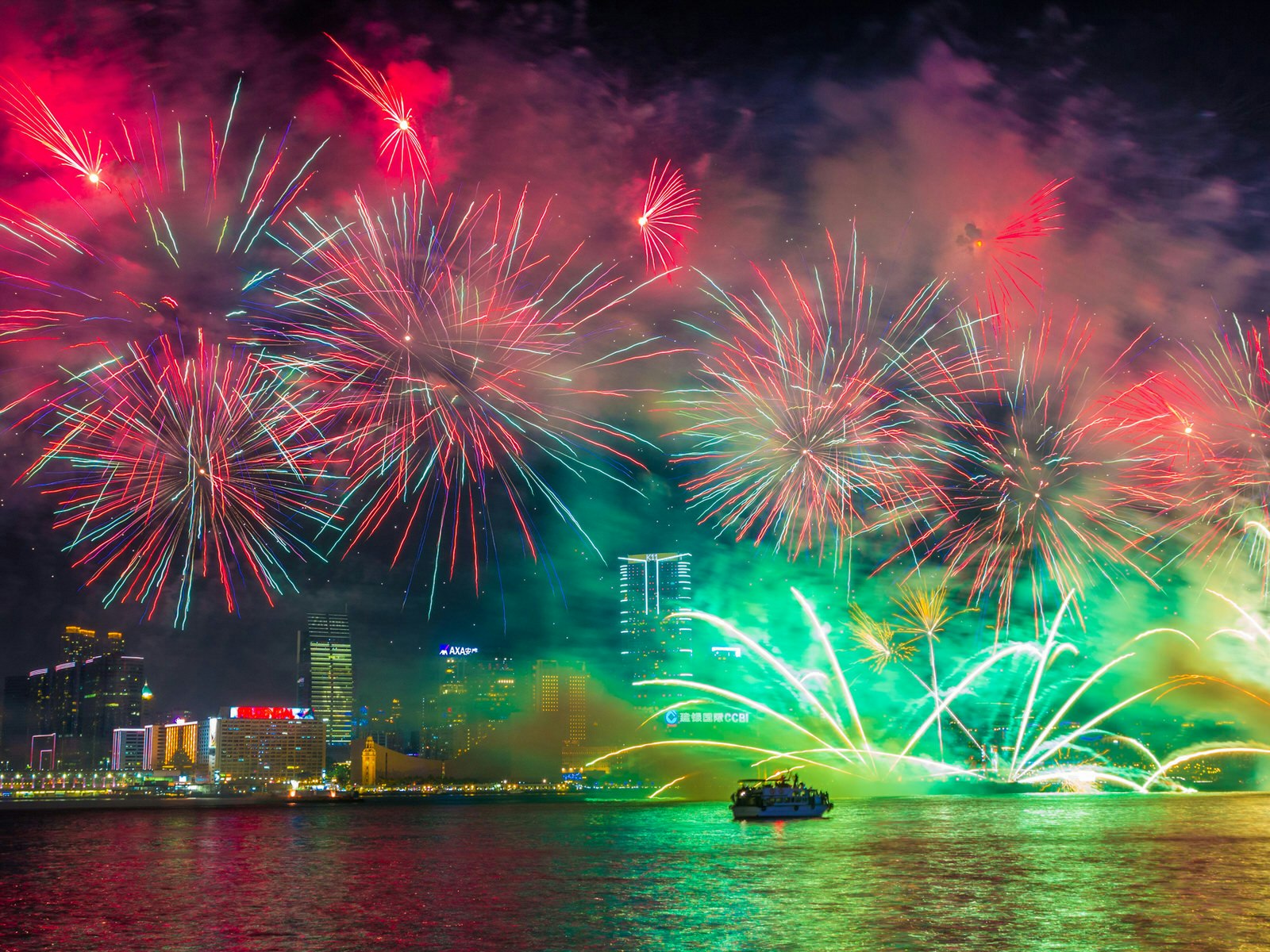 Red and green fireworks burst and reflect over the Hong Kong waterfront at night, with buildings lit up in the background and a boat in the foreground © e X p o s e / Shutterstock