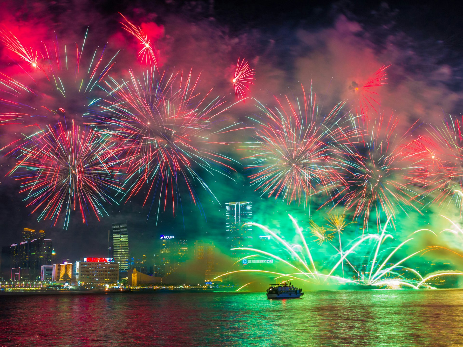 Red and green fireworks burst and reflect over the Hong Kong waterfront at night, with buildings lit up in the background and a boat in the foreground © e X p o s e / Shutterstock