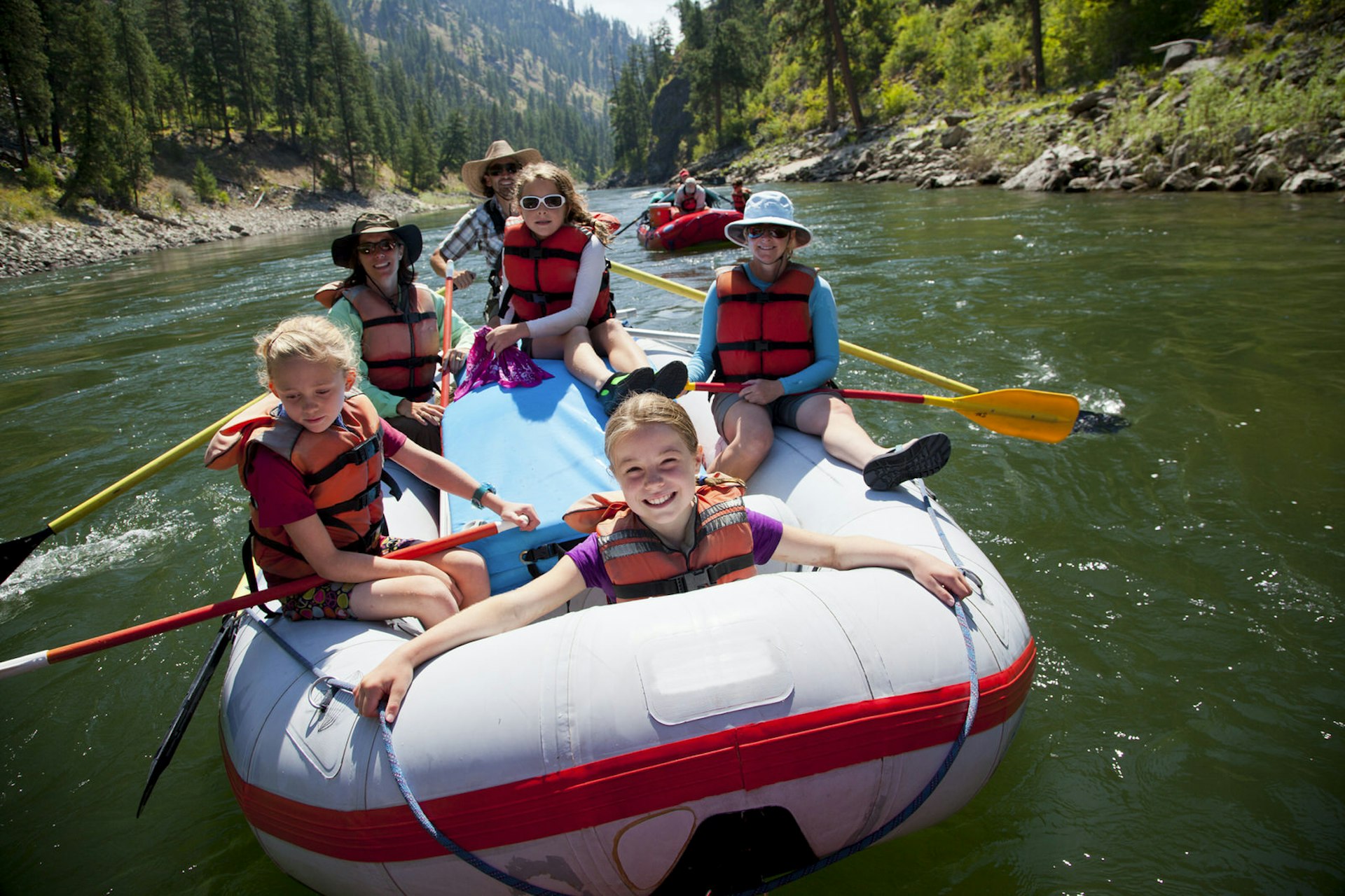 A group of kids smile as they ride in a whitewater raft down a green river in a canyon in Idaho.