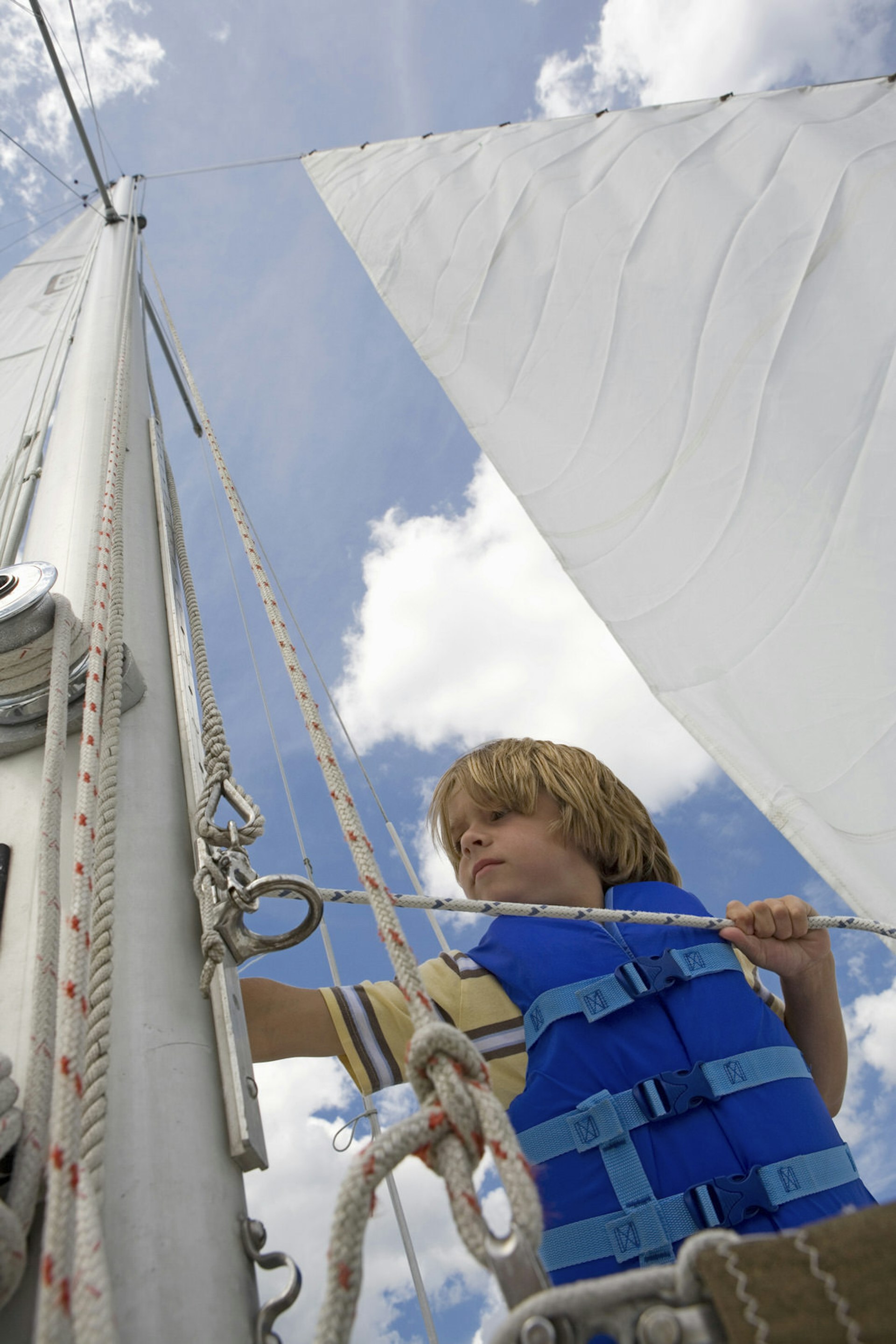A young boy wearing a bright blue life jacket ties a rope on a sailboat, with the sail towering above him