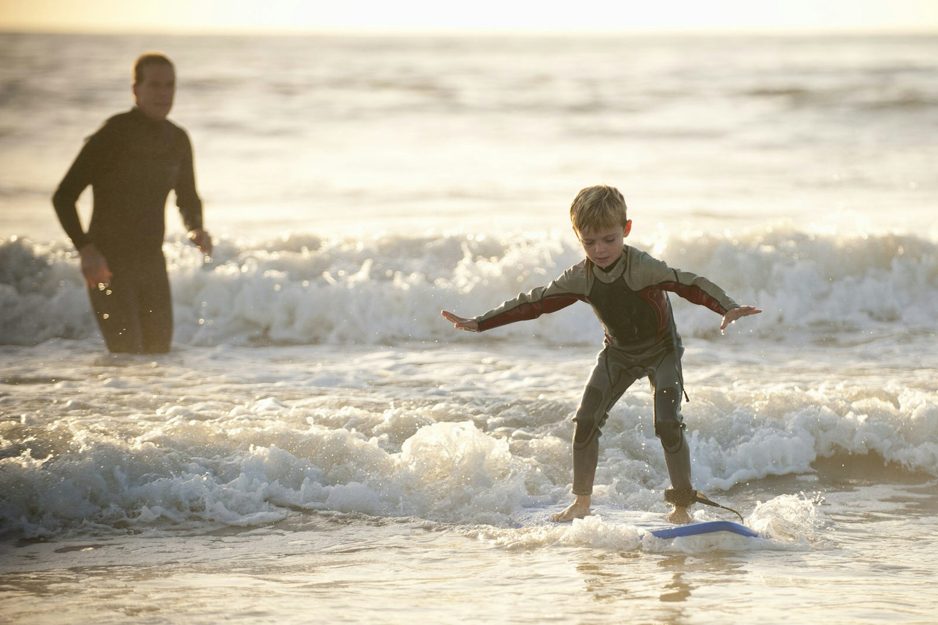 A father watches from a short distance as his small son learns to catch a wave while surfing close to the shoreline.