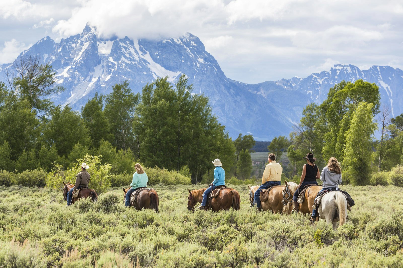 A group of horseback riders of all ages meanders through a meadow at the base of a mountain near Grand Teton National Park.