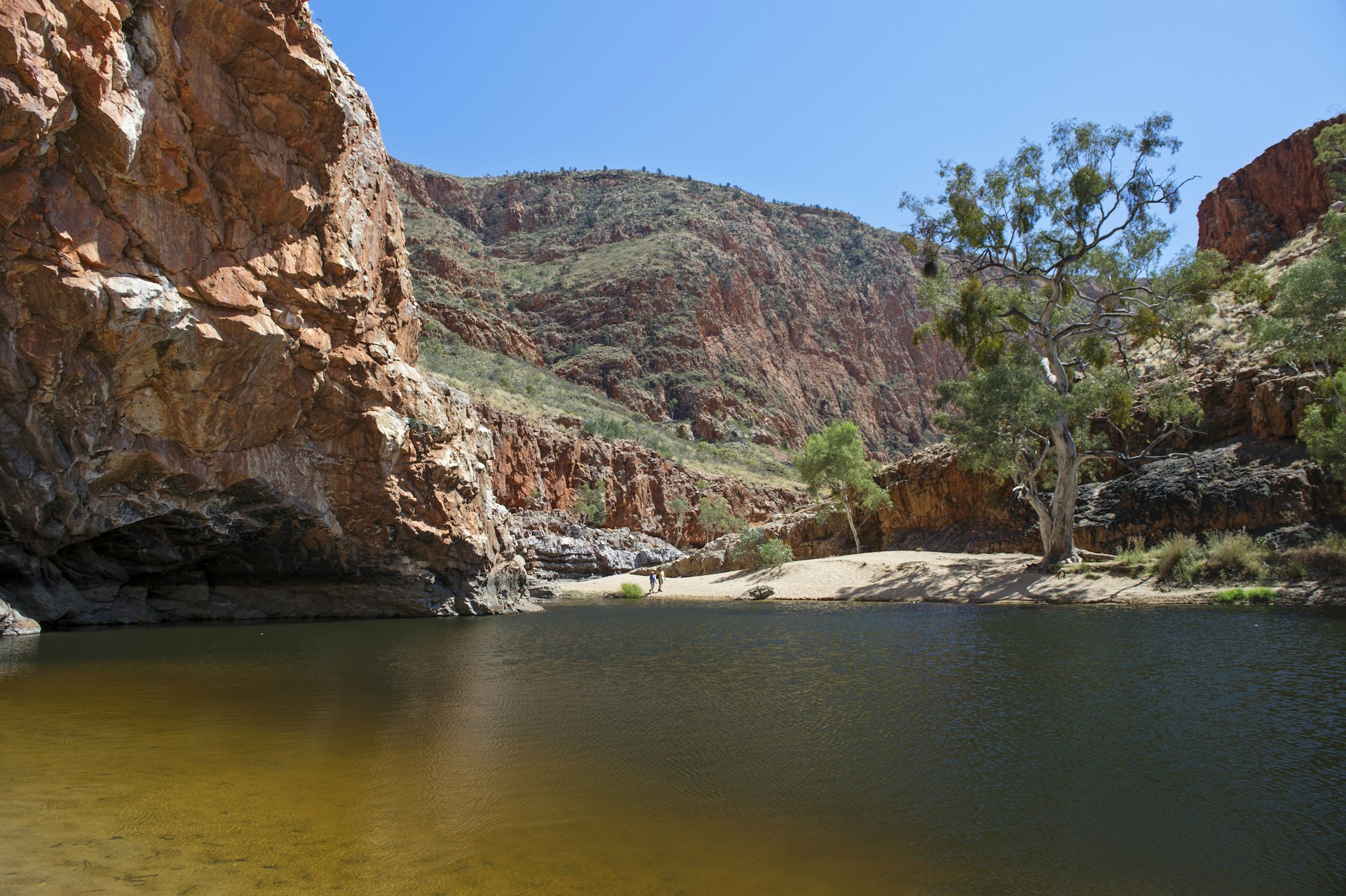 Ormiston Gorge, West MacDonnell National Park, Northern Territory. Pictured on a sunny day, the still body of water is surrounded by deep red sands and earth. Two people are walking away from the waters edge. There is blue sky and a few sparse trees in the background.