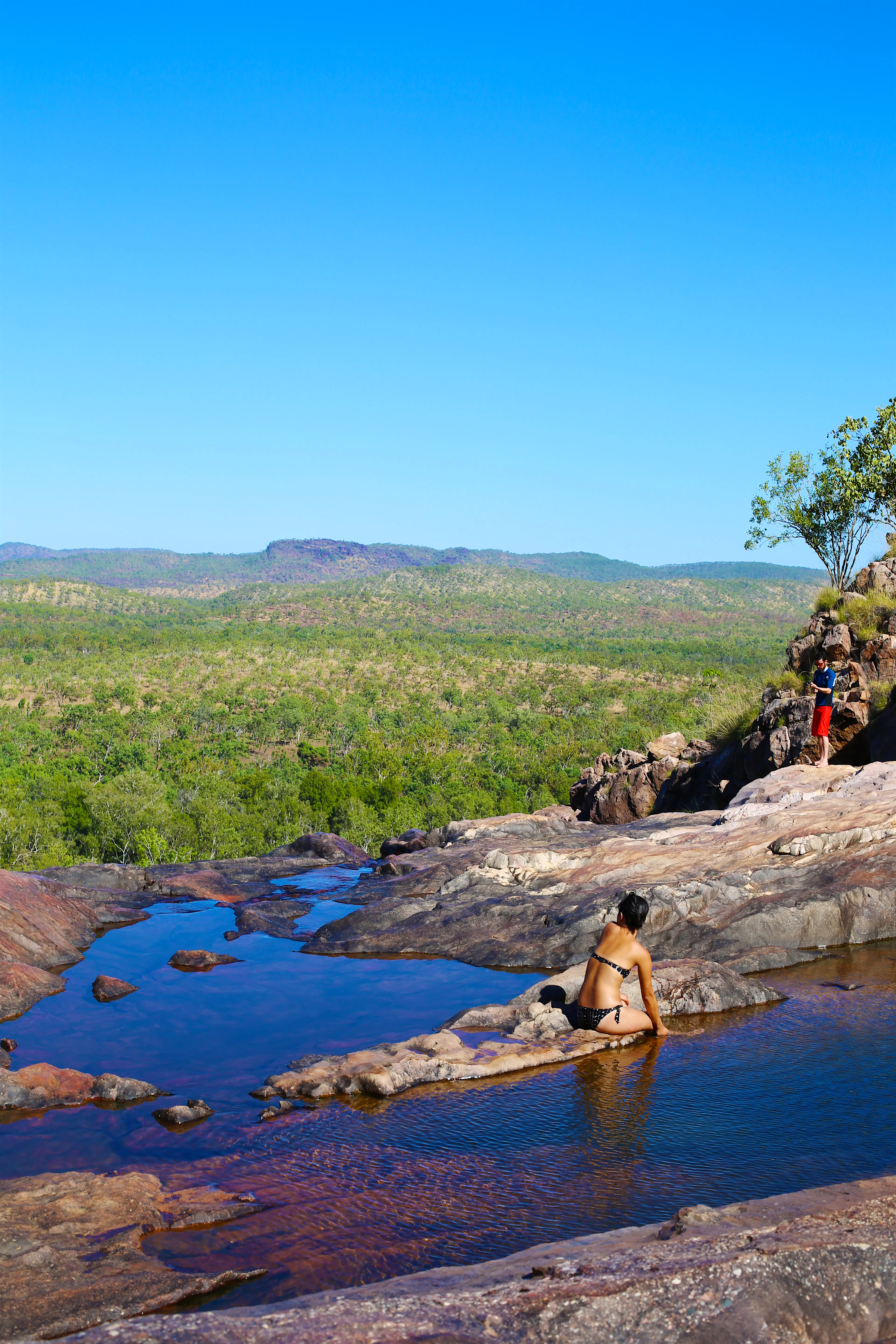 Features - Gunlom Falls, Kakadu National Park Gunlom Falls upper pools Northern Territory Australia. A woman sits an a small natural ledge between two pools wearing a black and white polka dot bikini, looking away from the camera. A man is standing in a pool further away, also looking away from the camera. It's a clear and sunny day