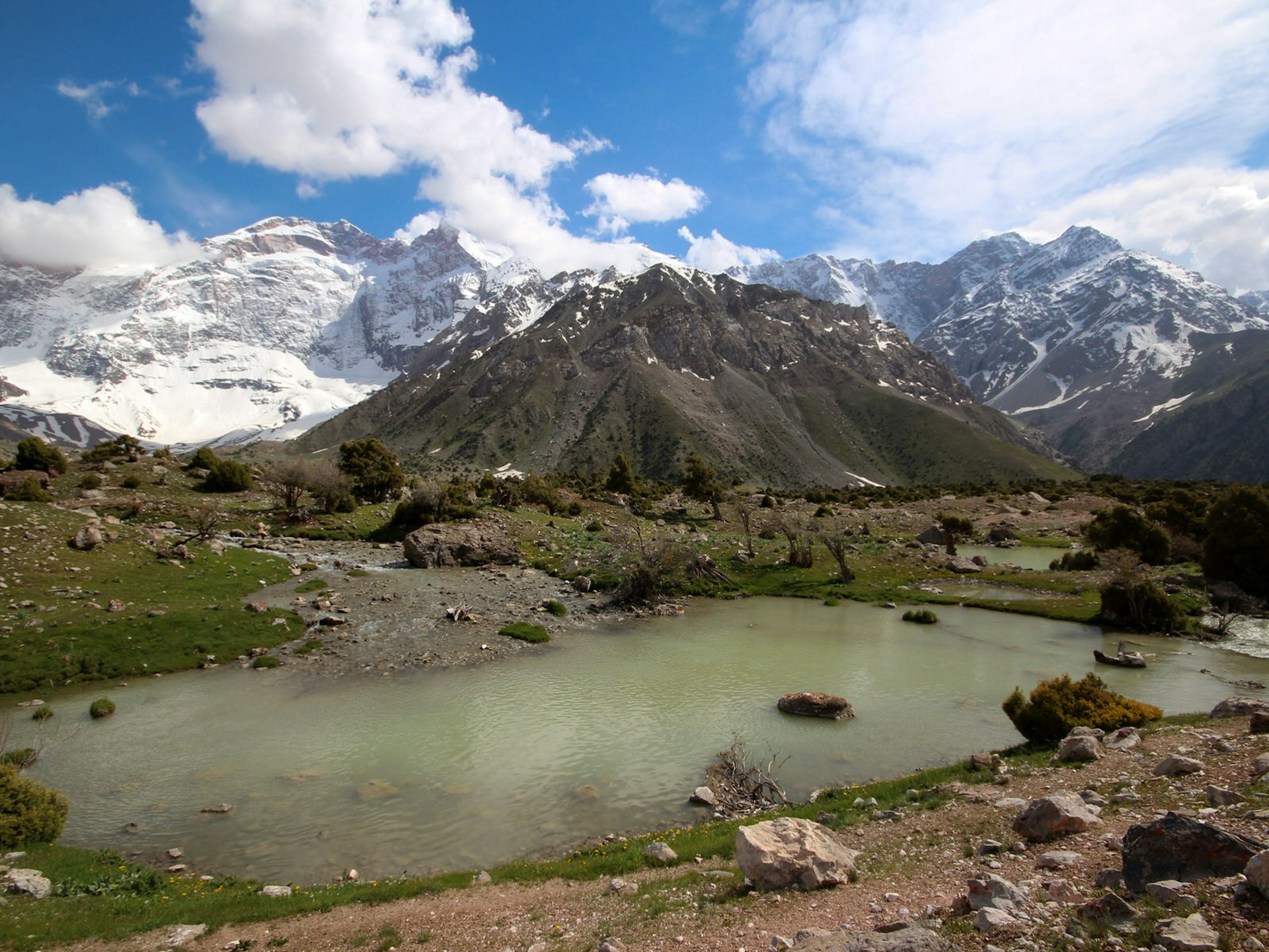 A small green lake in the foreground with snow-covered mountains and blue sky beyond. One of a dozen lakes in Tajikistan's Kulaikalon Basin © Stephen Lioy / Lonely Planet