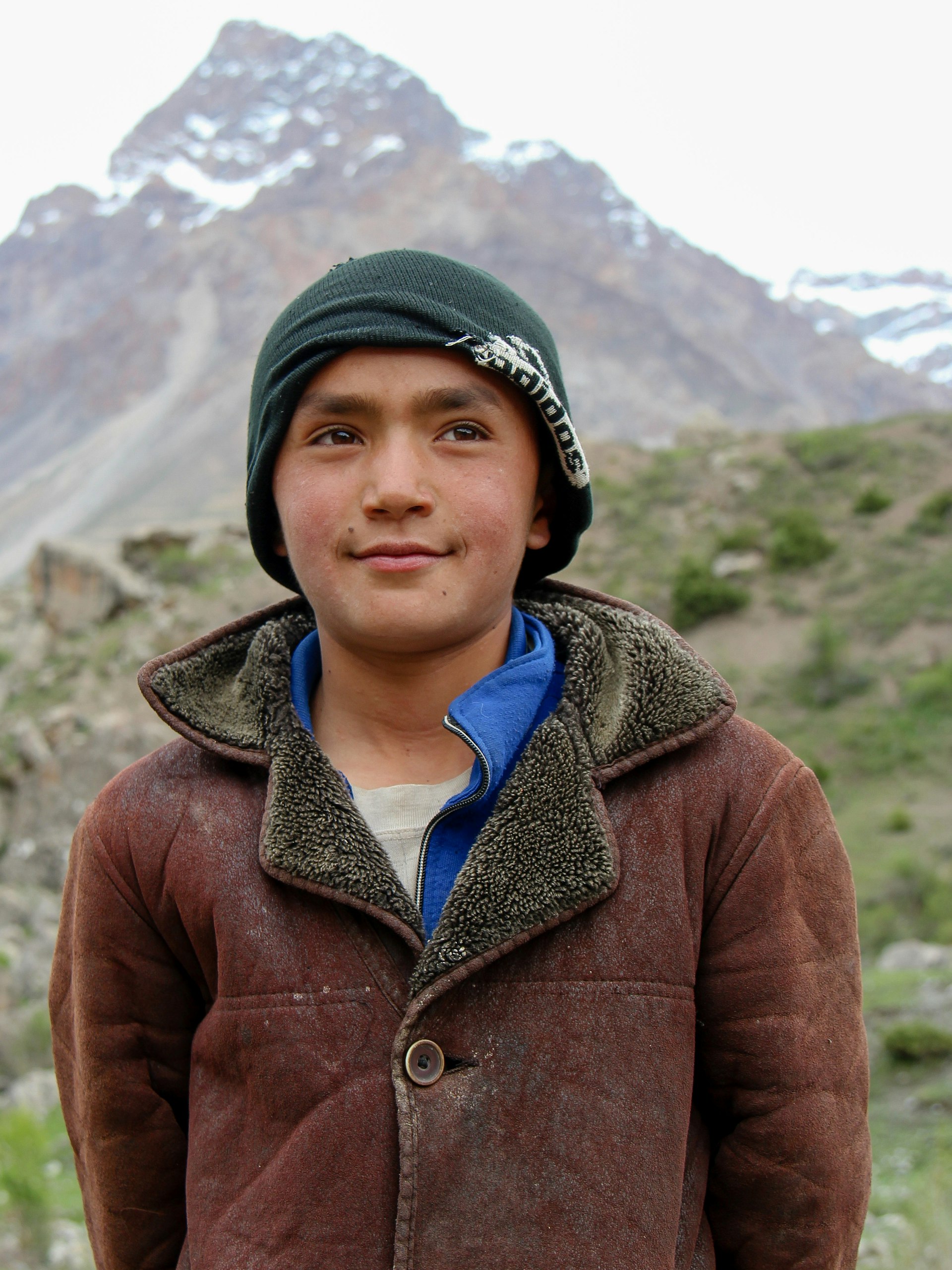 A young Tajik man dressed in a brown suede coat and green head scarf stands in front of a snowy mountain peak © Stephen Lioy / Lonely Planet