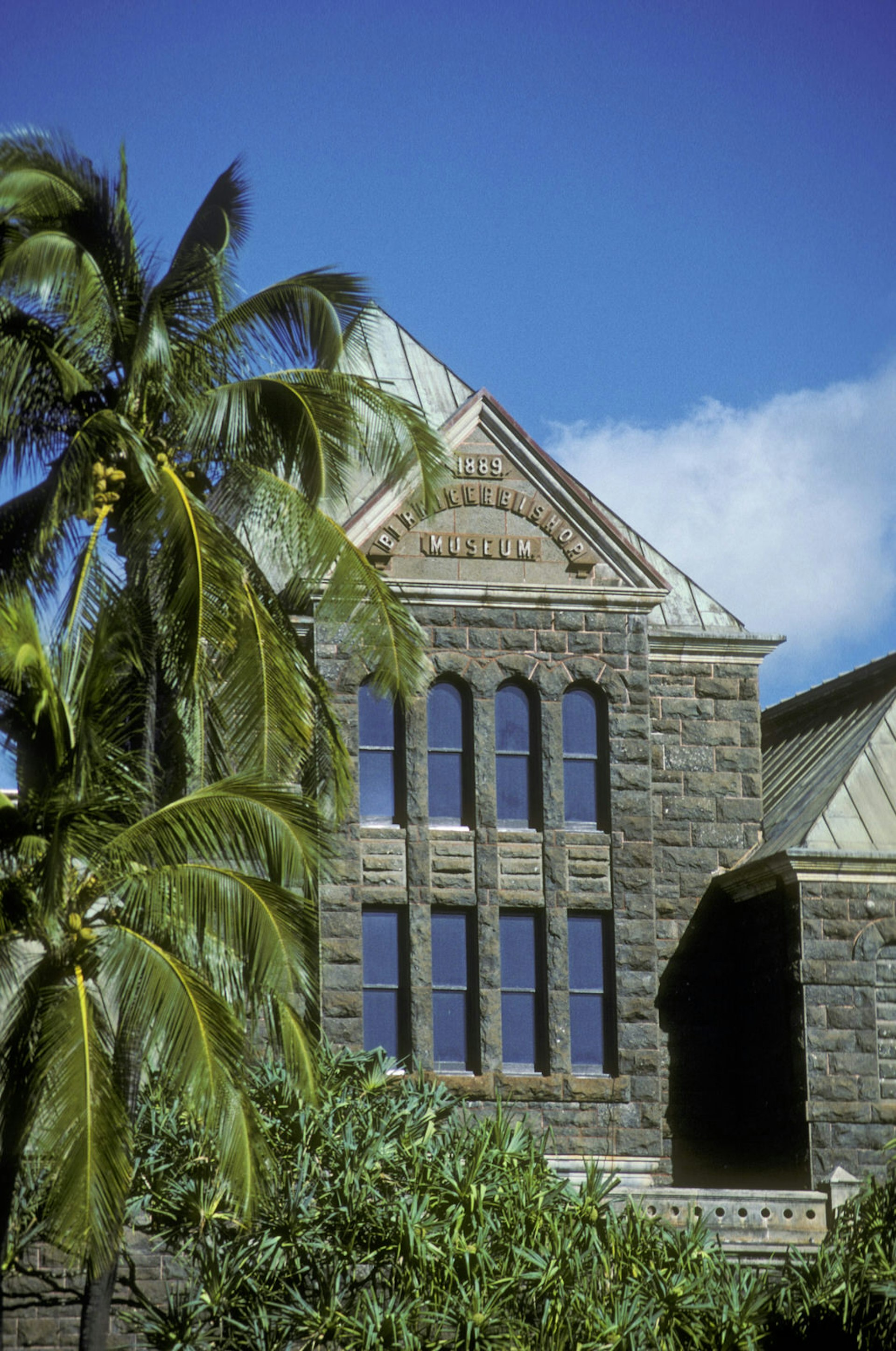 One wing of the Bishop Museum is seen in the foreground, with a gray stone facade. Palm trees in the foreground © Greg Vaughn / Getty Images