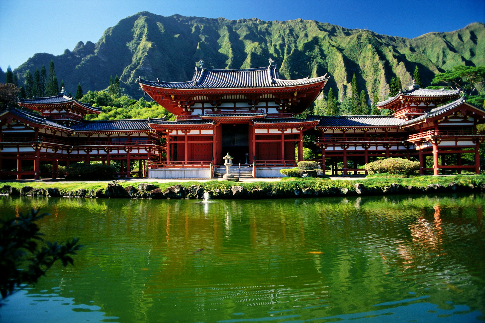 A large japanese-style temple, with red wooden columns and walls, sits at the bottom of a green mountain, and is reflected in the green water of a pond in the foreground © Van de Ven Mary / Getty Images