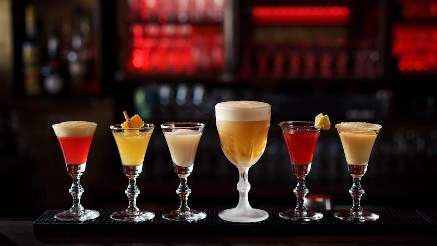 A selection of drinks on a dark bar top. The drinks are served in small glasses with bevelled stems and are a range of colours with a range of garnishes on top. 