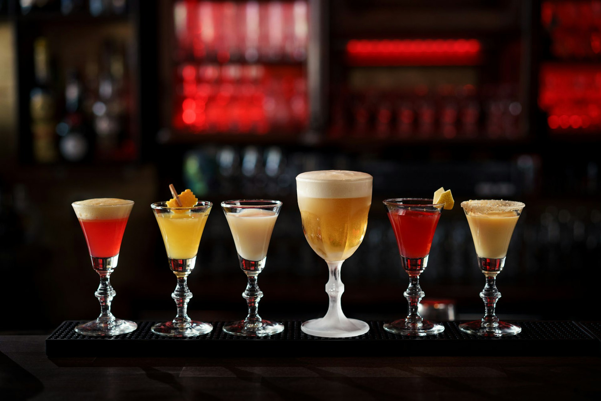 A selection of drinks on a dark bar top. The drinks are served in small glasses with bevelled stems and are a range of colours with a range of garnishes on top. 
