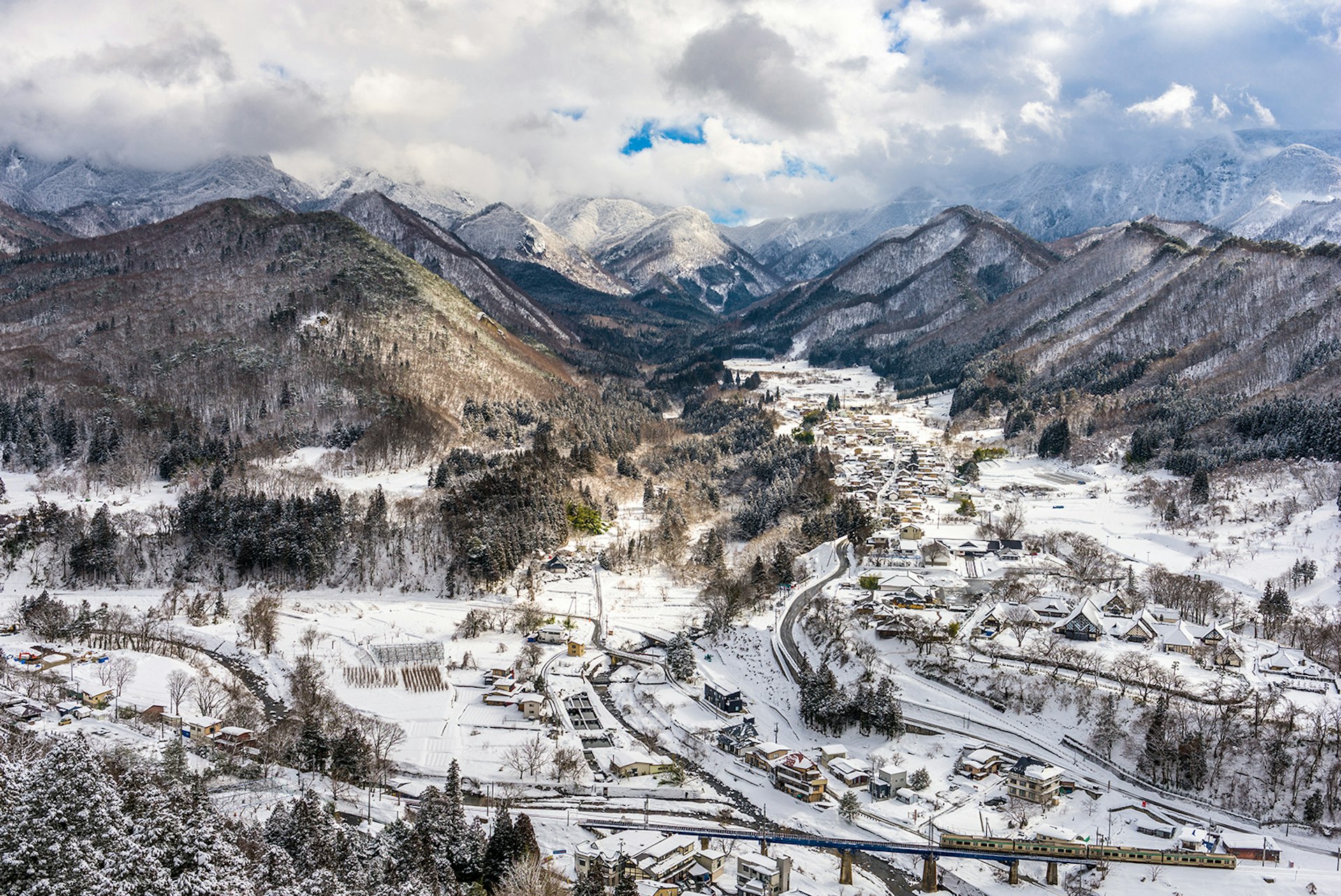 An aerial view of a snow-covered valley with a small village. A mountain range stretches off into the distance.