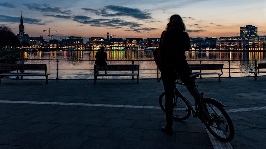 See Hamburg in a weekend by bike. A photo of a cyclist overlooking the canal at dusk. We can see one of Hamburg's famous spires silhouetted against the colourful sky, the lights from the buildings are reflected in the water. 