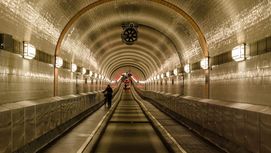 An image taken inside the Old Elbe Tunnel, Hamburg. The domed tunnel is tiled with white and black tiles and illuminated by old-fashioned lights every few metres. someone is wheeling their bike through the tunnel and in the far distance we can see red brake lights. 