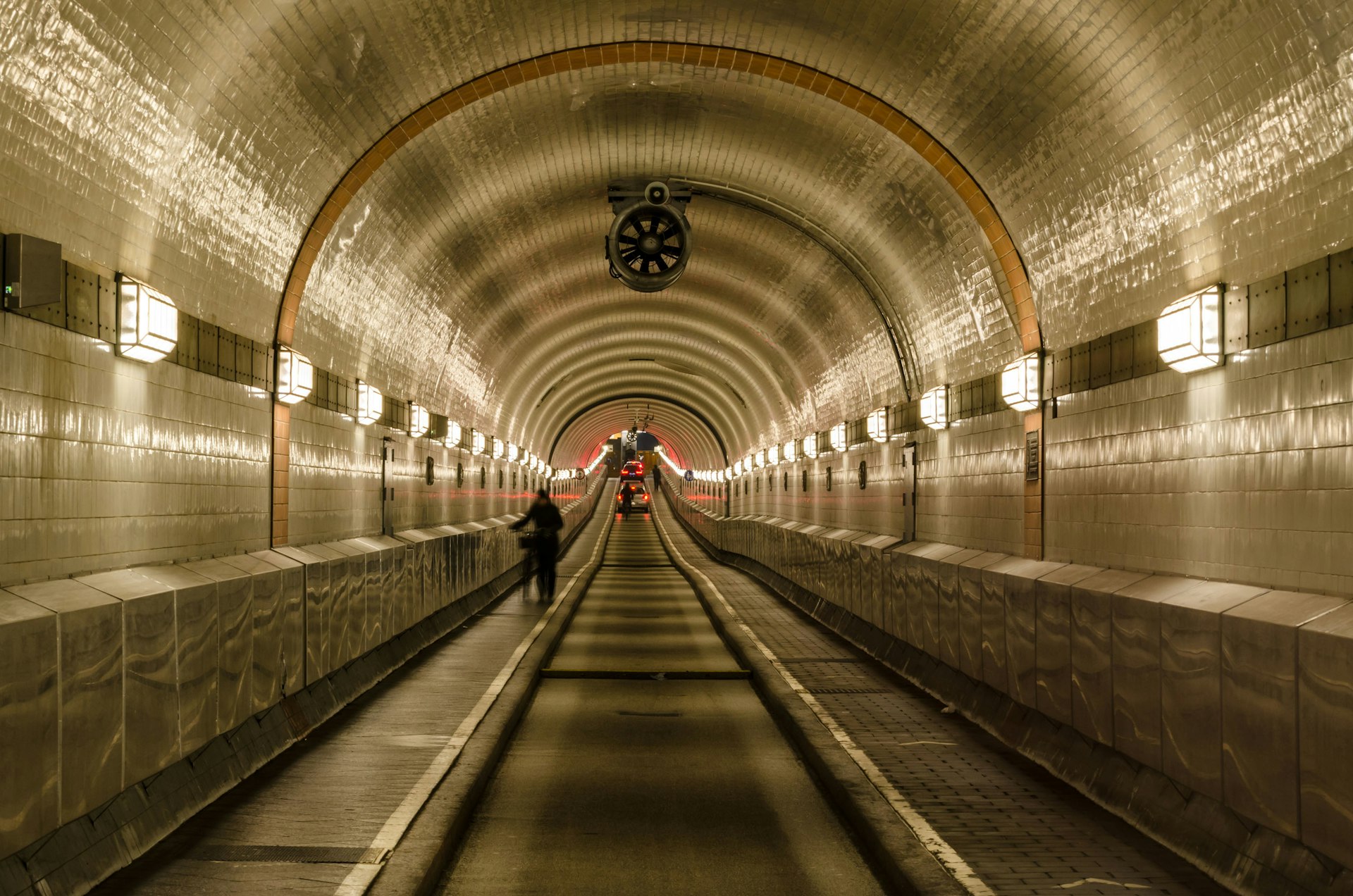 An image taken inside the Old Elbe Tunnel, Hamburg. The domed tunnel is tiled with white and black tiles and illuminated by old-fashioned lights every few metres. someone is wheeling their bike through the tunnel and in the far distance we can see red brake lights. 