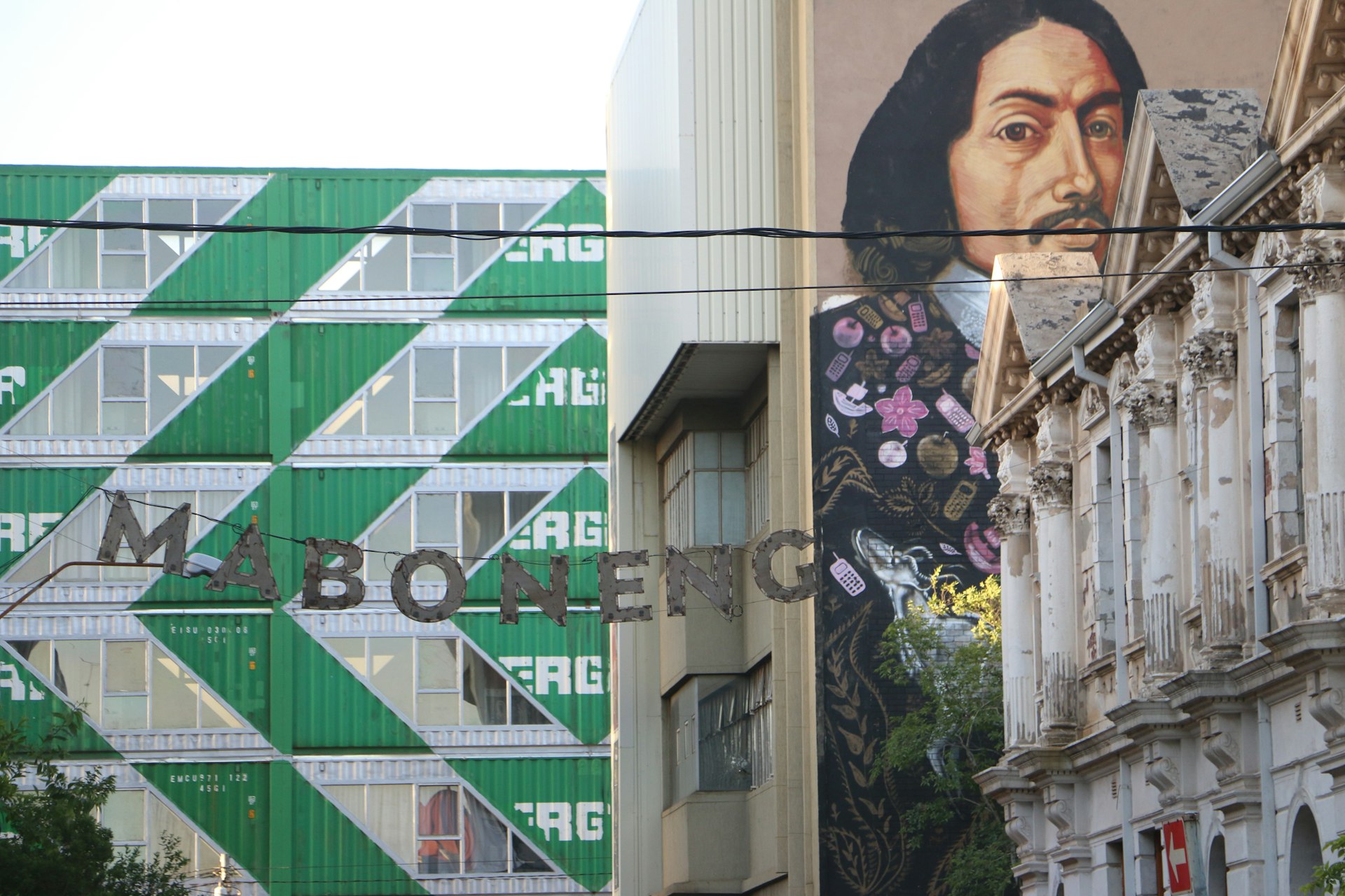 A set of metal letters spelling Maboneng hang across a street in Johannesburg. Wealthered colonial buildings stand to the right, in front of a three-storey tall mural of Jan Van Riebeeck. A tower of stacked green-and-white shipping containers, with windows cut out, stands in the background © Simon Richmond / Lonely Planet