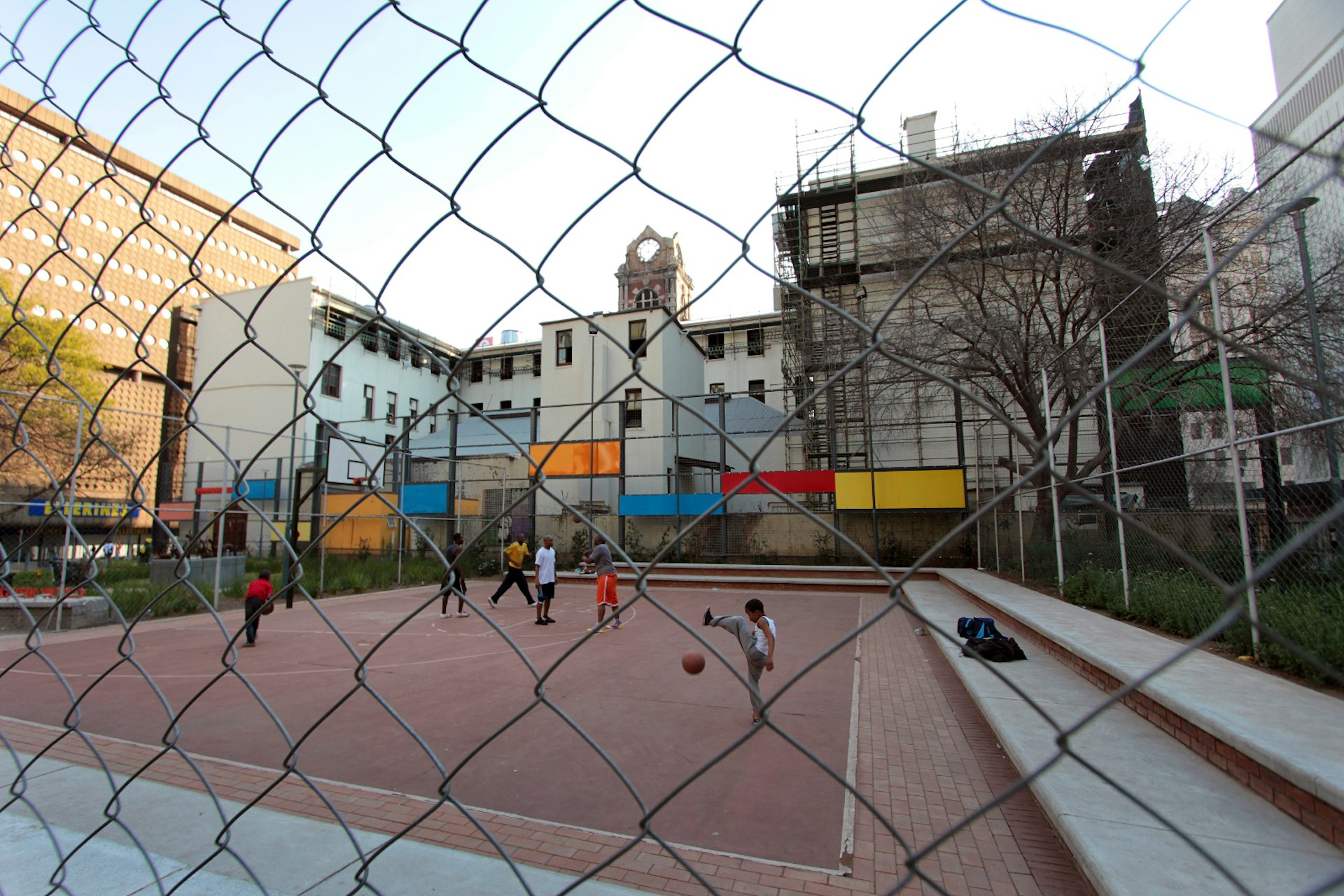 Looking through the chainlinked fence at the public basektball court in Ernest Oppenheimer Park, with a group in action - a young boy in the foreground tries to bounce the ball between his legs © Heather Mason / Lonely Planet