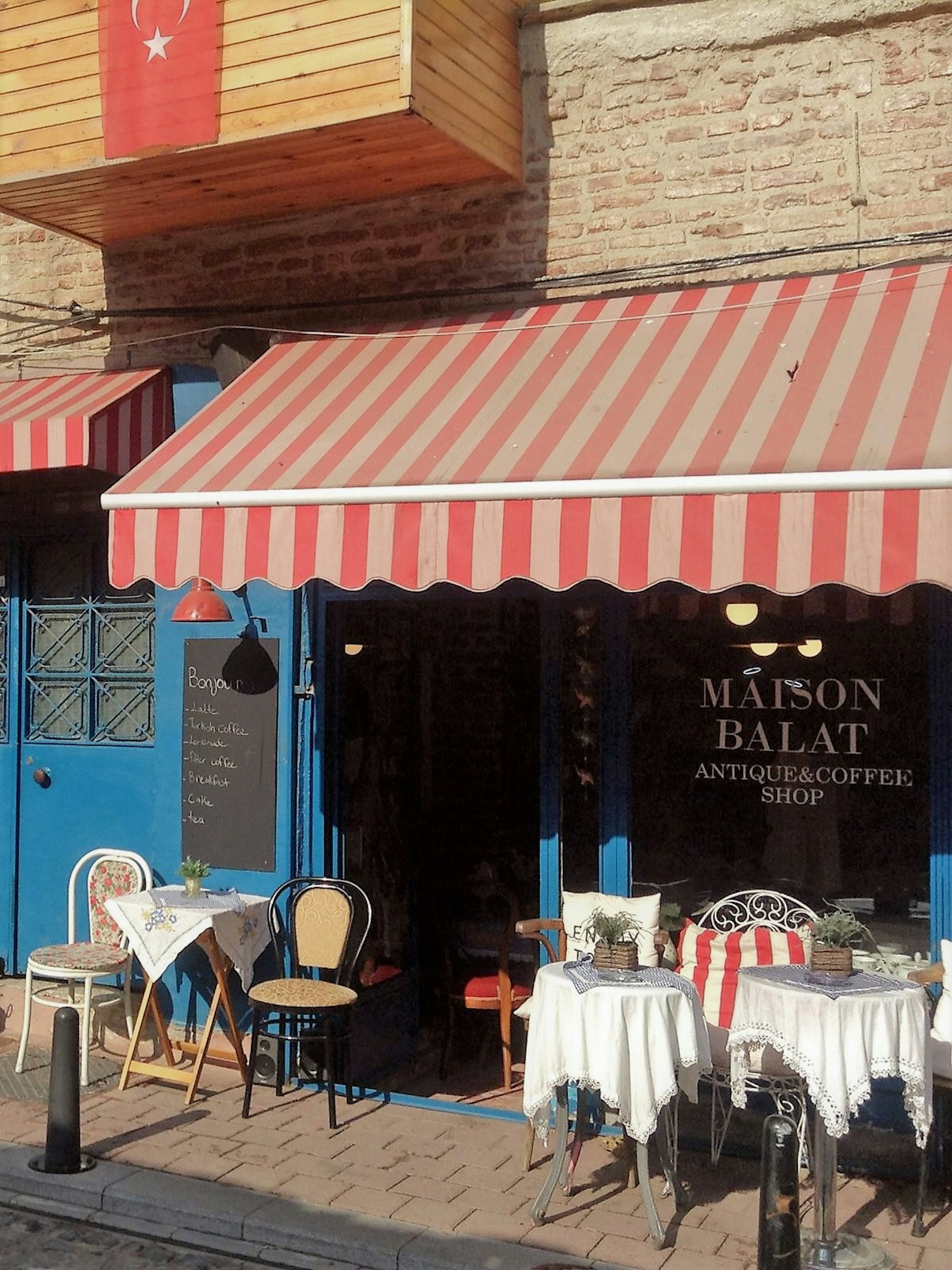 A french cafe and antiques shop with a vibrant, primary colour blue facade and a red and white striped awning. Mismatched chairs and table with delicate tablecloths sit outside on a sunny day in front of the glass shopfront window displaying the name 'Maison Balat' © Jennifer Hattam / Lonely Planet