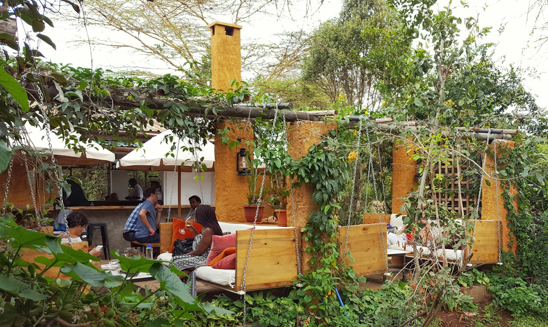 Wooden chairs with large white cushions hang from chains in a very leafy outdoor garden. Patrons relax while sipping coffee © Clementine Logan / Lonely Planet