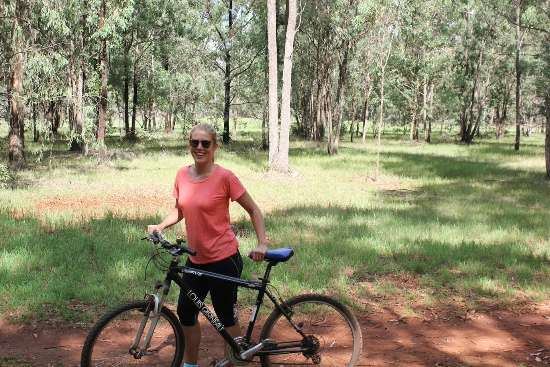Writer Clementine Logan stands smiling behind her mountain bike on a red dirt trail within Kakura Forest, Nairobi© Clementine Logan / Lonely Planet