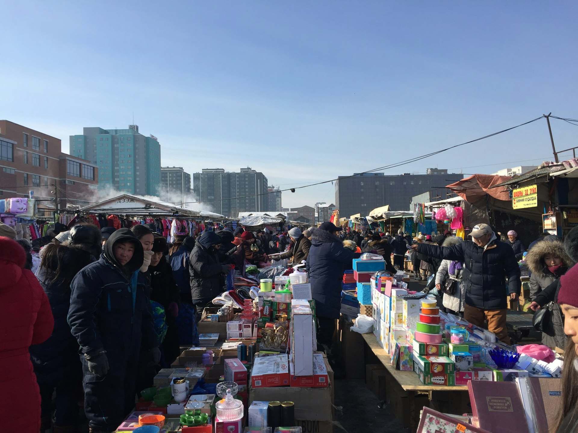 Naran Tuul Market: an outdoor market with rows of tables filled with products for sale and packed with shoppers in big coats, under a blue sky