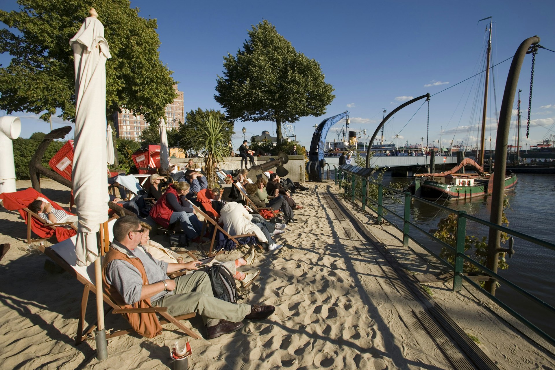 People sitting on deckchairs on a manmade beach on the banks of the Elbe river, enjoying a weekend in Hamburg. It is a sunny day and there are white umbrellas alongside the chairs. 