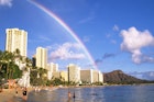 People wade through the waters off Waikiki beach, as a long clear rainbow arches its way to diamond head in the distance.