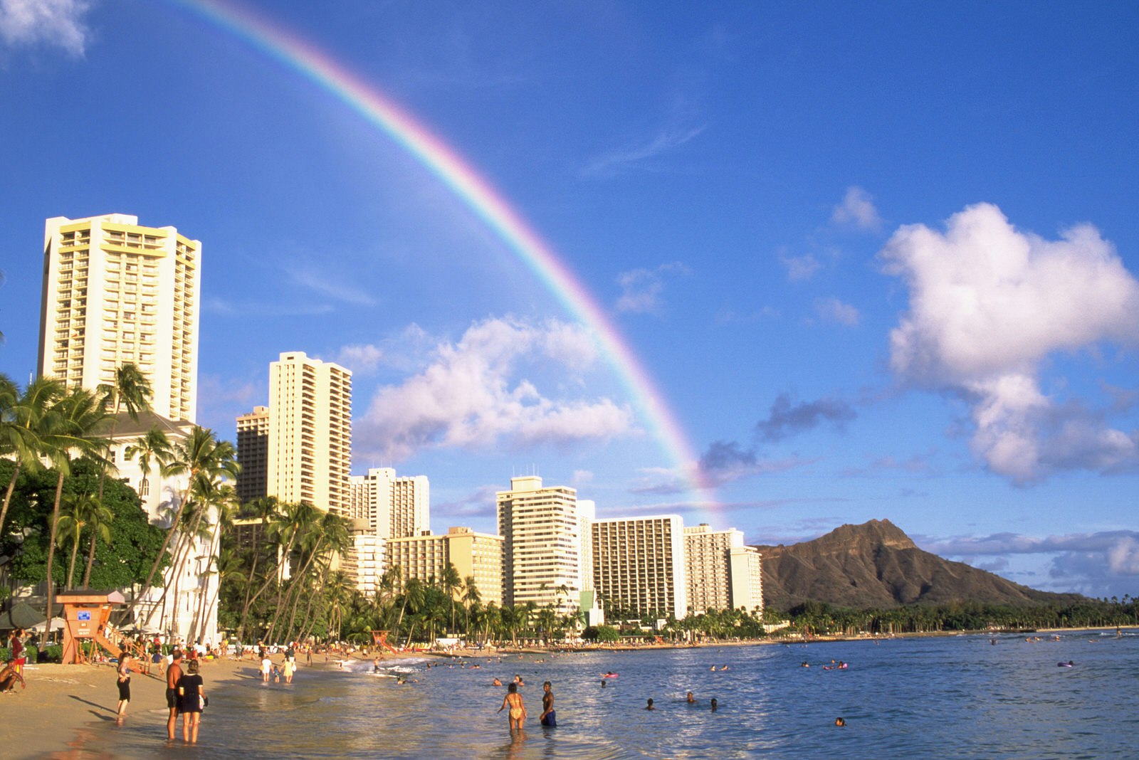 People wade through the waters off Waikiki beach, as a long clear rainbow arches its way to diamond head in the distance © Elfi Kluck / Getty Images