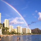 People wade through the waters off Waikiki beach, as a long clear rainbow arches its way to diamond head in the distance.