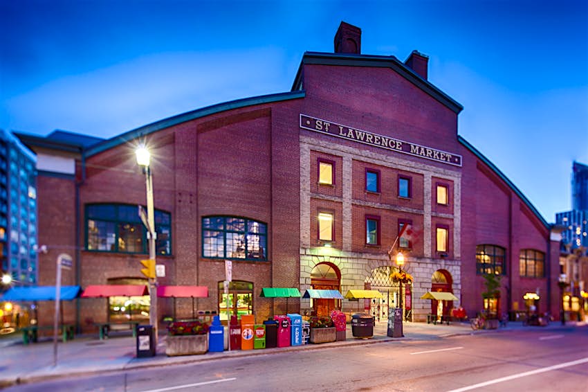 Lights glow at twilight around the giant brick facade of the St. Lawrence Market, one of two major markets in Toronto.