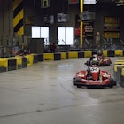 Yellow and black barriers border the wide expanse of a twisting go-kart track, where riders in red cars speed around a corner.