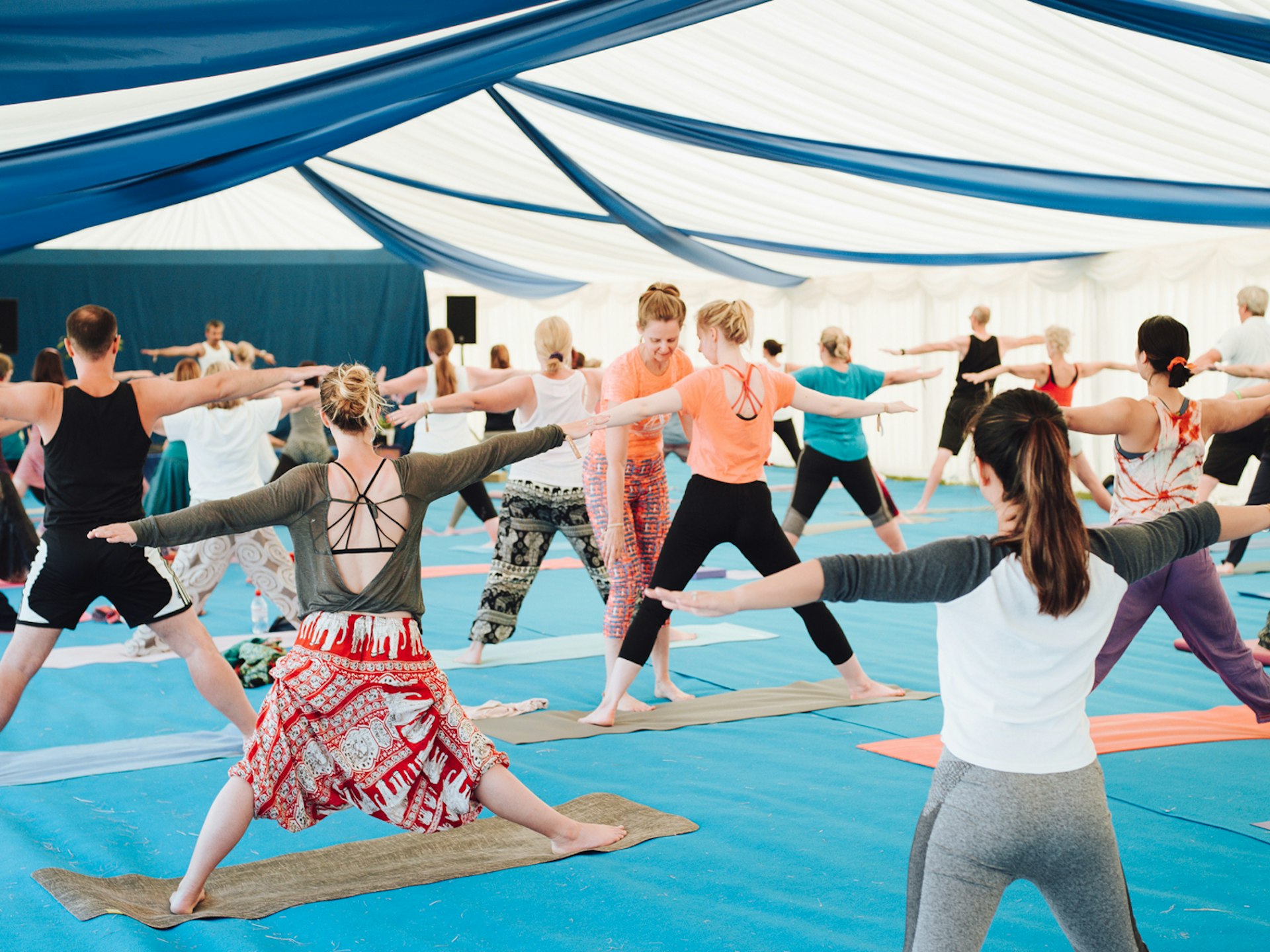 Alternative festivals - Lots of participants enjoying a yoga class inside a large tent with a white and blue draped material roof. The floor is blue and the yogis are about to enter the triangle pose. An instructor is making corrections to one person's posture.