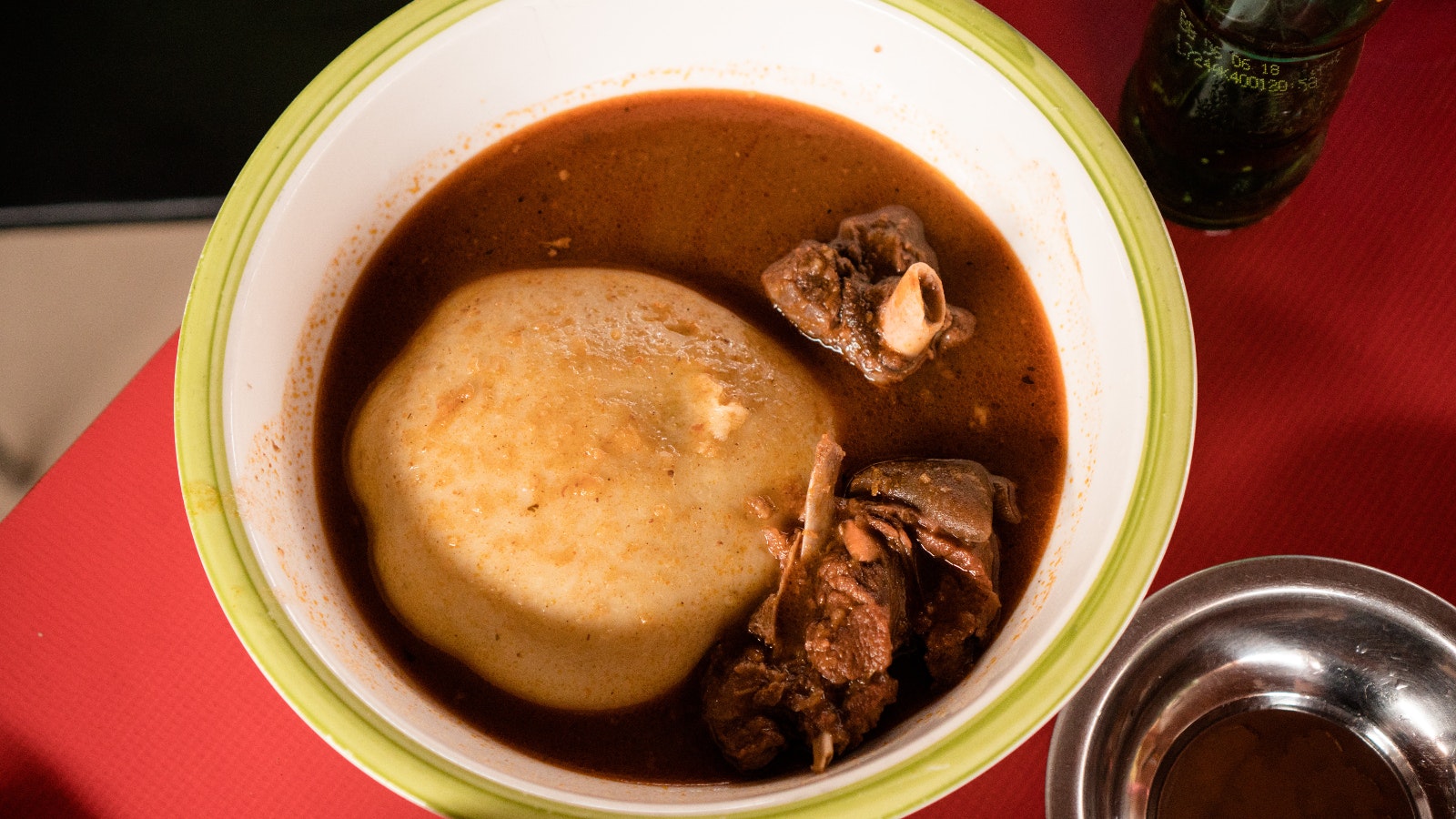 Within a green-rimmed white bowl (on a red table cloth) is the traditional dish of fufu. Sitting in reddish light soup is a ball of yellowish dough made from cooked (and pounded) plantain and cassava. Next to this sits some goat meat and bones © Elio Stamm / Lonely Planet