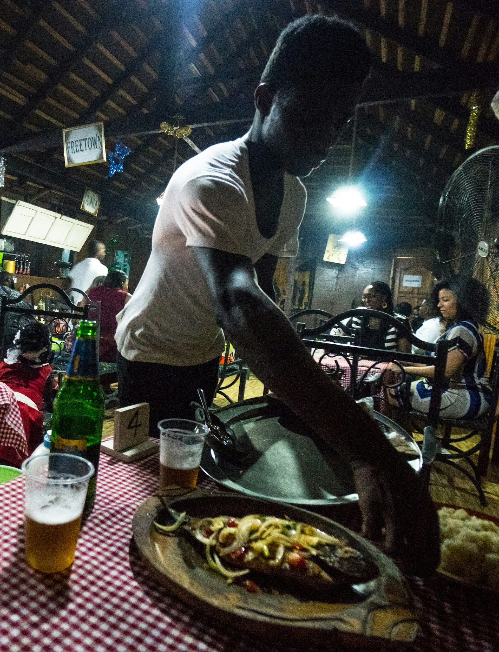 Floodlit, yet feeling dark, the confines of Chez Clarisse Mama Africa in Accra. A waiter in a white t-shirt leans over the red-and-white chequered tablecloth to place a dish of fried tilapia on the table © Elio Stamm / Lonely Planet