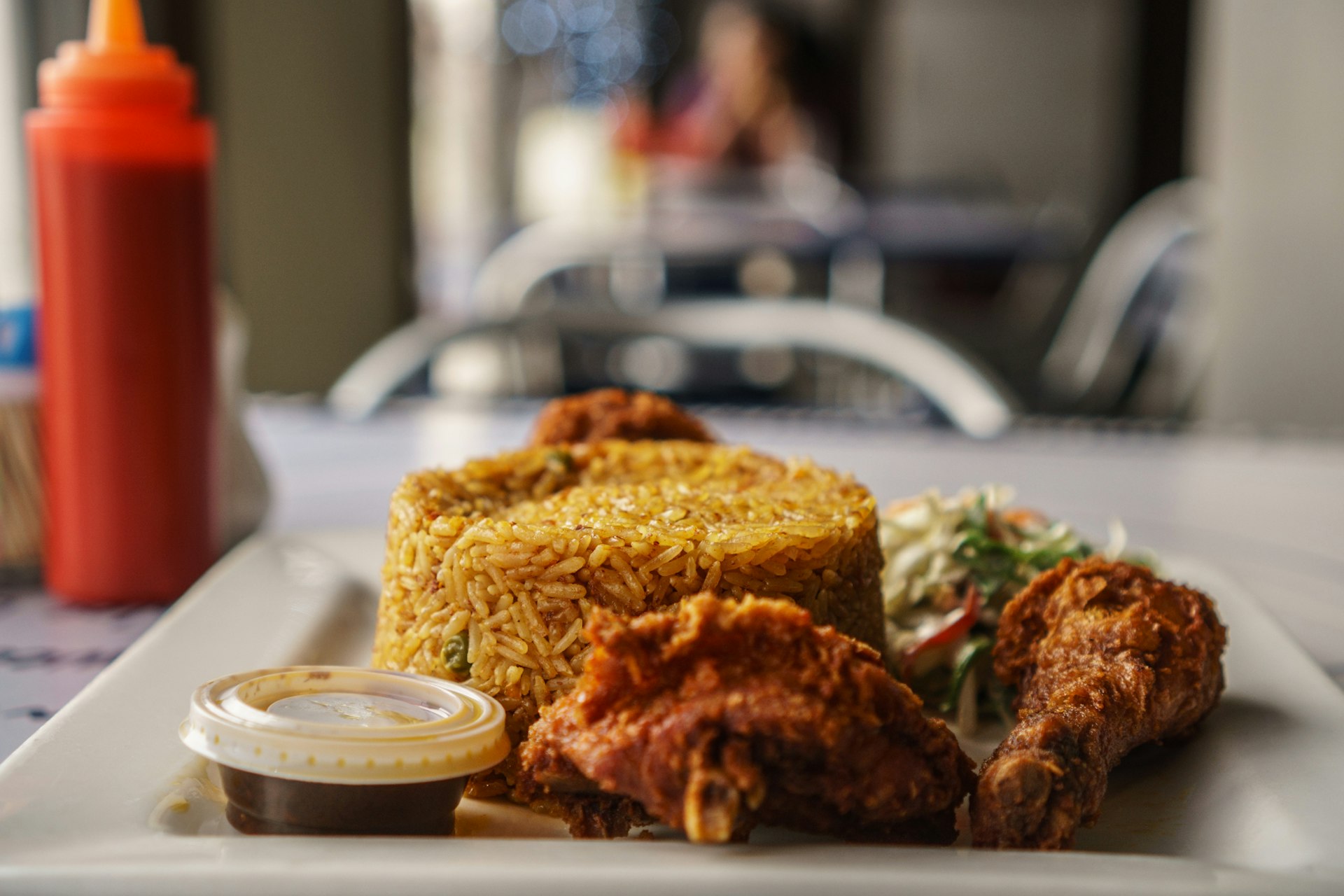  Flanked by a bottle of tomato ketchup, a square plate is laden with a neat cylindrical pile of yellow Jollof rice, two fried chicken legs, a salad and small plastic container of chilli sauce. Taken at Frankie's © Elio Stamm / Lonely Planet