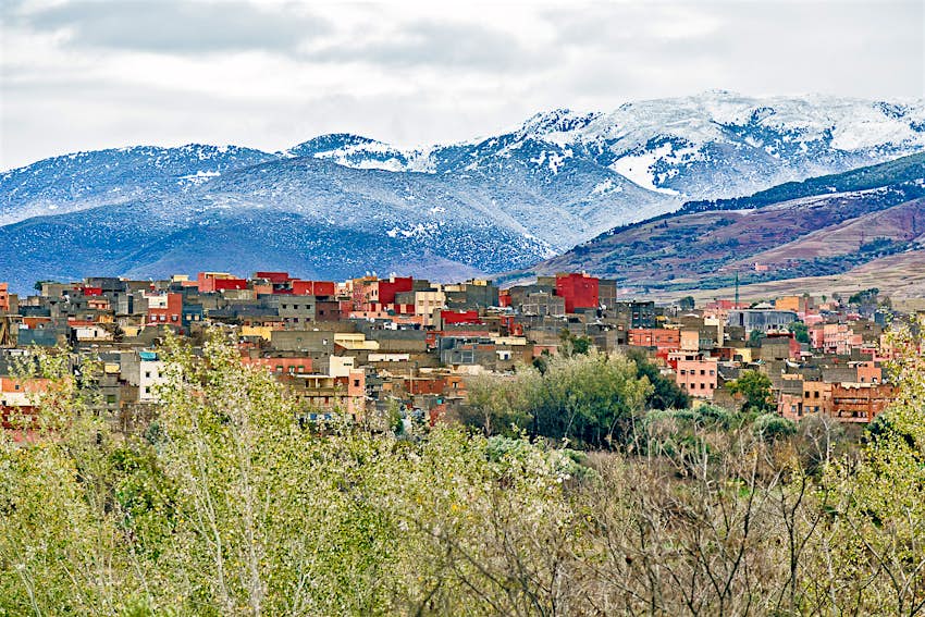 Amizmiz, a small village of atlas in Marrakesh, Morocco. Yellow and terracotta coloured square buildings sit behind green leafy trees. The snow-capped Atlas mountains are in the background, beneath a cloudy, grey sky. 