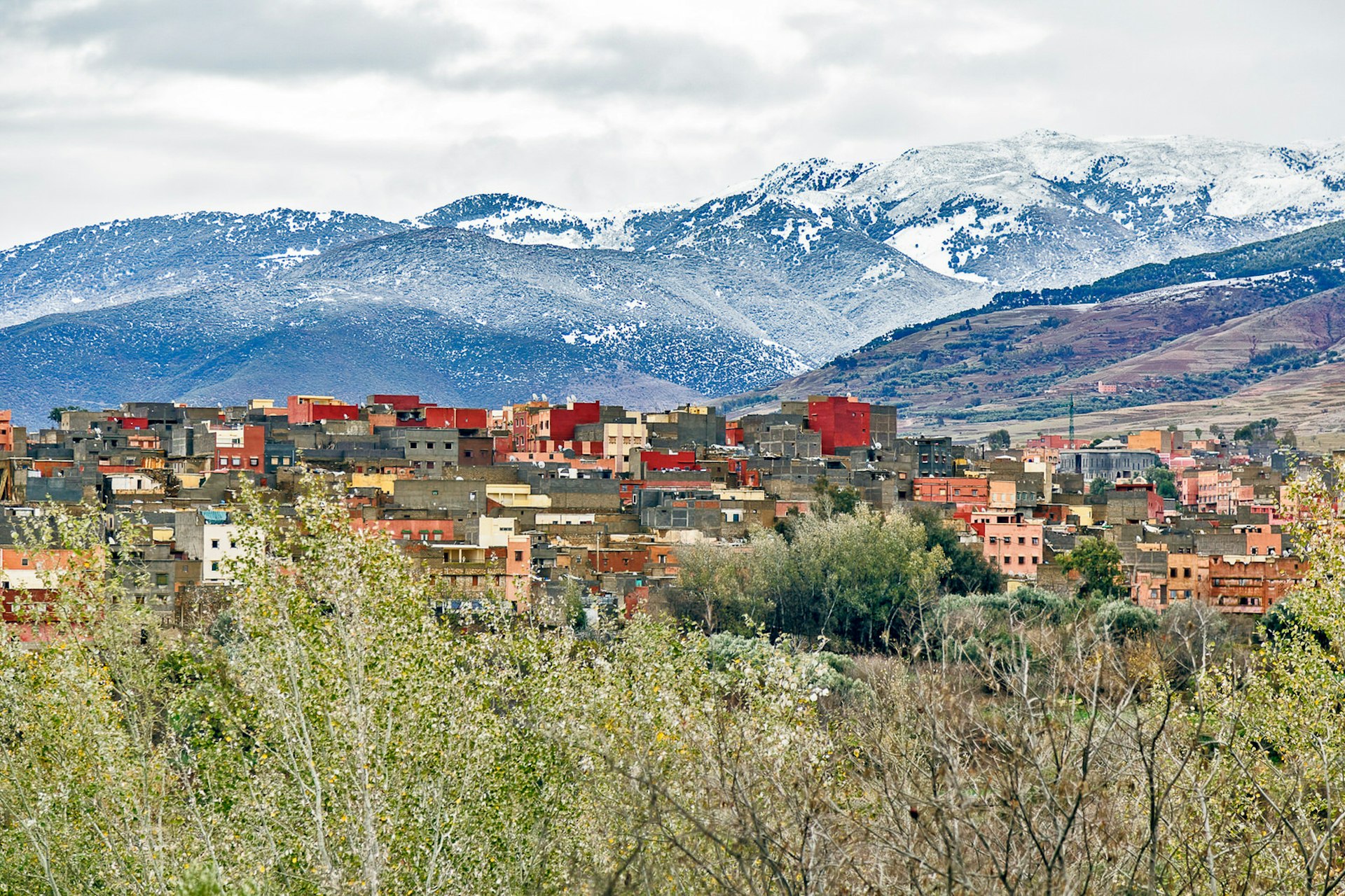 Amizmiz, a small village of atlas in Marrakesh, Morocco. Yellow and terracotta coloured square buildings sit behind green leafy trees. The snow-capped Atlas mountains are in the background, beneath a cloudy, grey sky. 