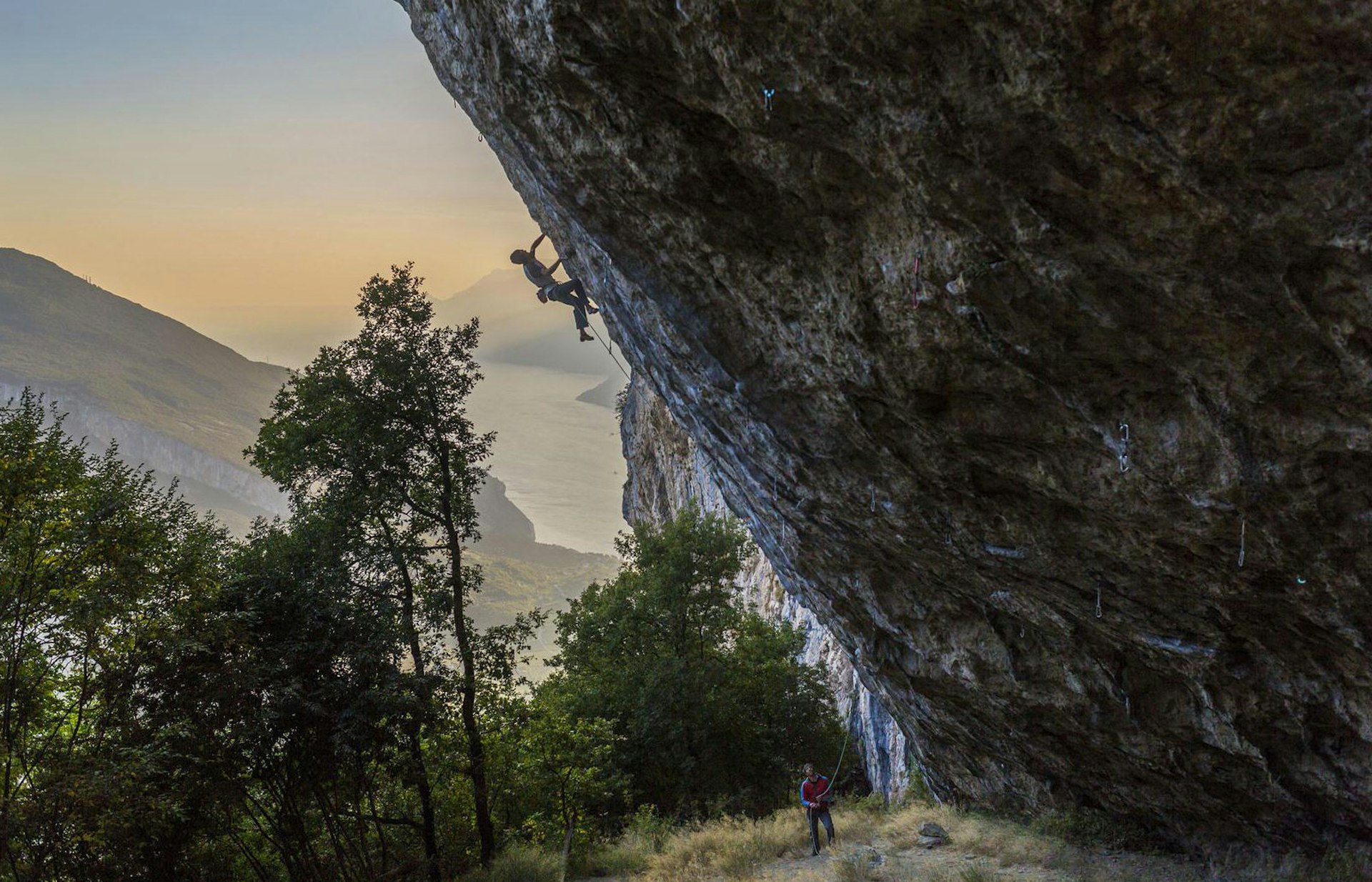 A climber scales an overhanging rockface, with Lake Garda in the distance