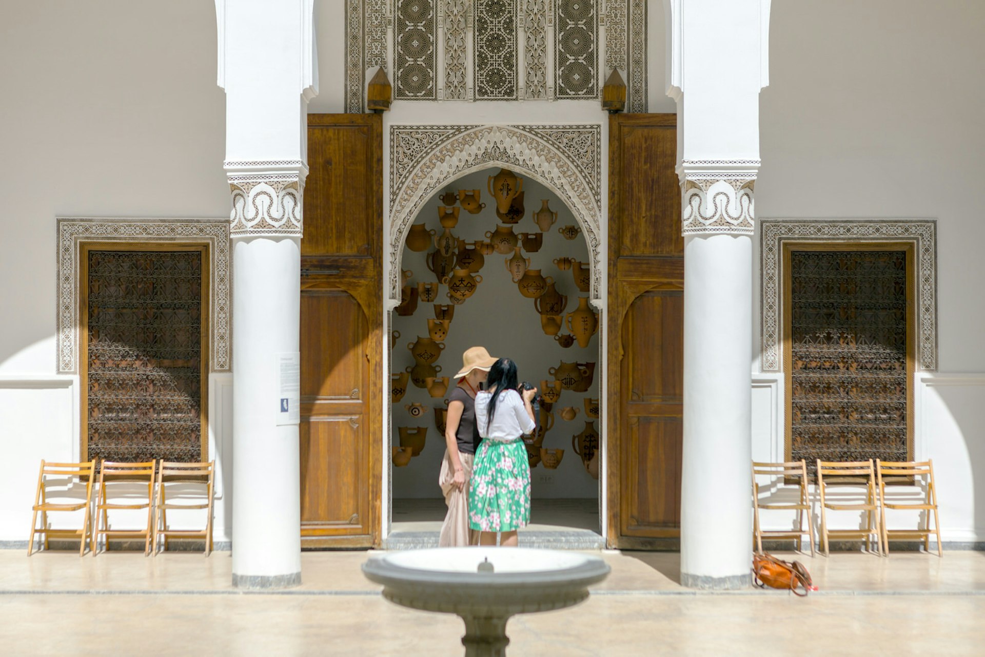 Courtyard of Dar Bellarj, Marrakesh, Morocco. A small stone fountain is in the foreground, behind it two women look at a camera, framed by the archway of a tall decorated door. Through the door it appears that clay teapots are suspended in the air. 