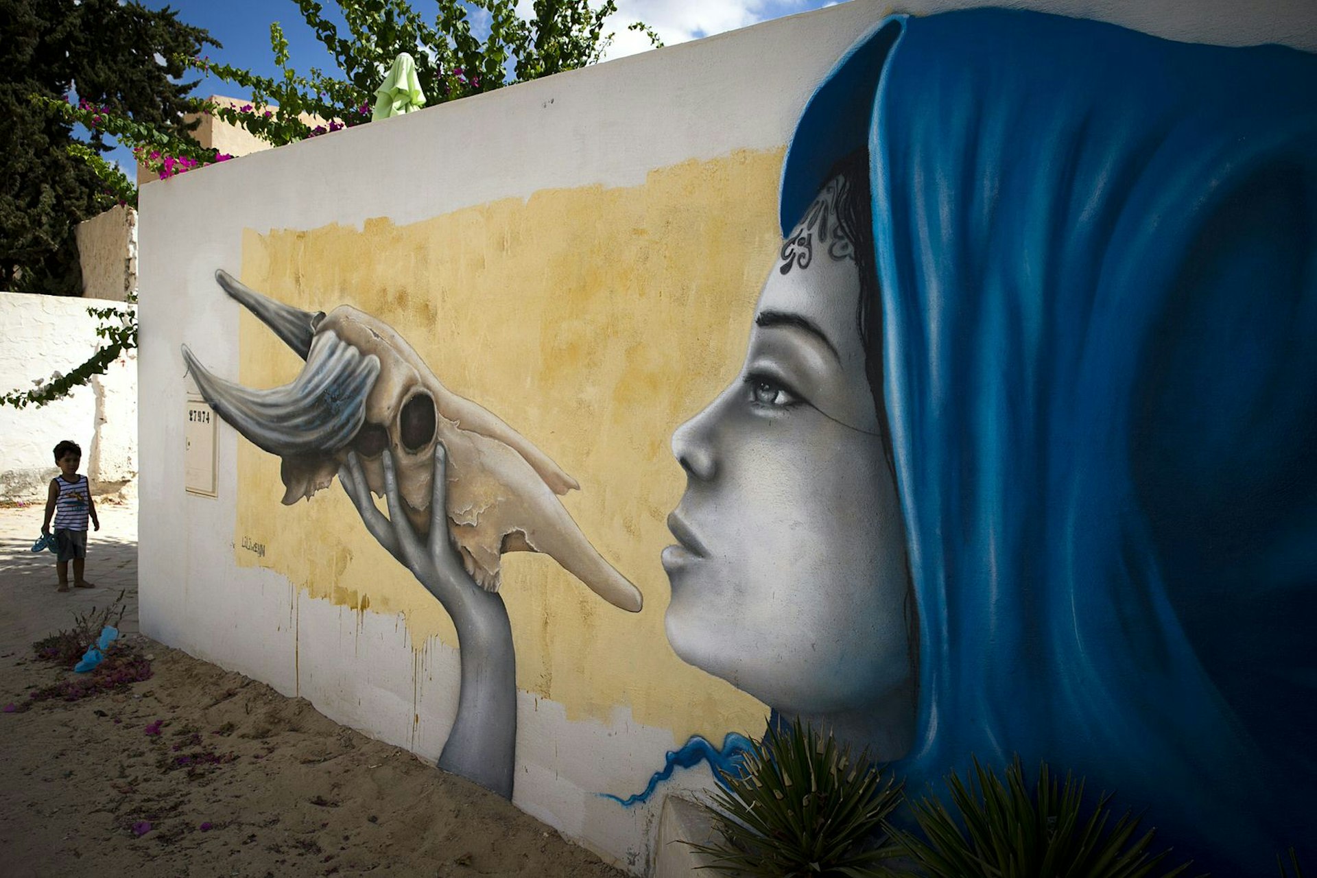 A child stands by a mural by French artist LILIWENN which decorates a wall in the village of Erriadh, on the Tunisian island of Djerba, on August 8, 2014, as part of the artistic project "Djerbahood". Nearly 100 artists from 30 different nationalities were invited by French-Tunisian organizer Mehdi Ben Cheikh to take part in an initiative to turn Djerba's Erriadh town into an "open sky museum". The village of Erriadh is one of the oldest in Tunisia where Jews, Muslims and Christians have lived together for centuries.
