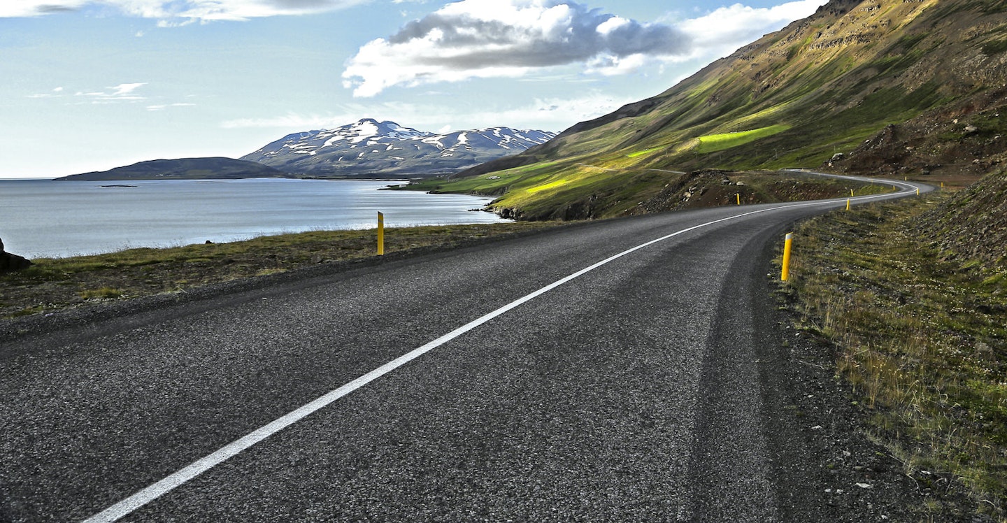 curving-road-cuts-through-green-landscape-on-edge-of-eyjafjordur-with-snow-capped-mountains-in-distance