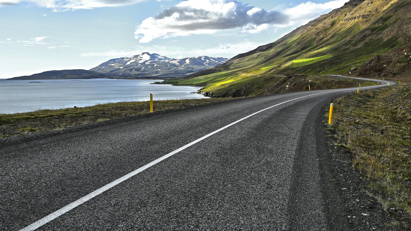curving-road-cuts-through-green-landscape-on-edge-of-eyjafjordur-with-snow-capped-mountains-in-distance