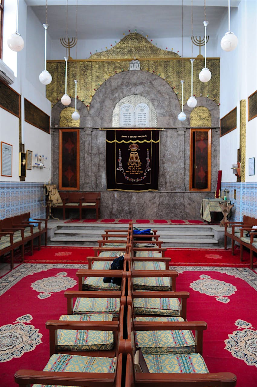 An inside view of the Lazama Synagogue, the oldest synagogue in the old Jewish quarter of Marrakesh, Morocco. Cushioned, wooden chairs sit on patterned red carpets. 