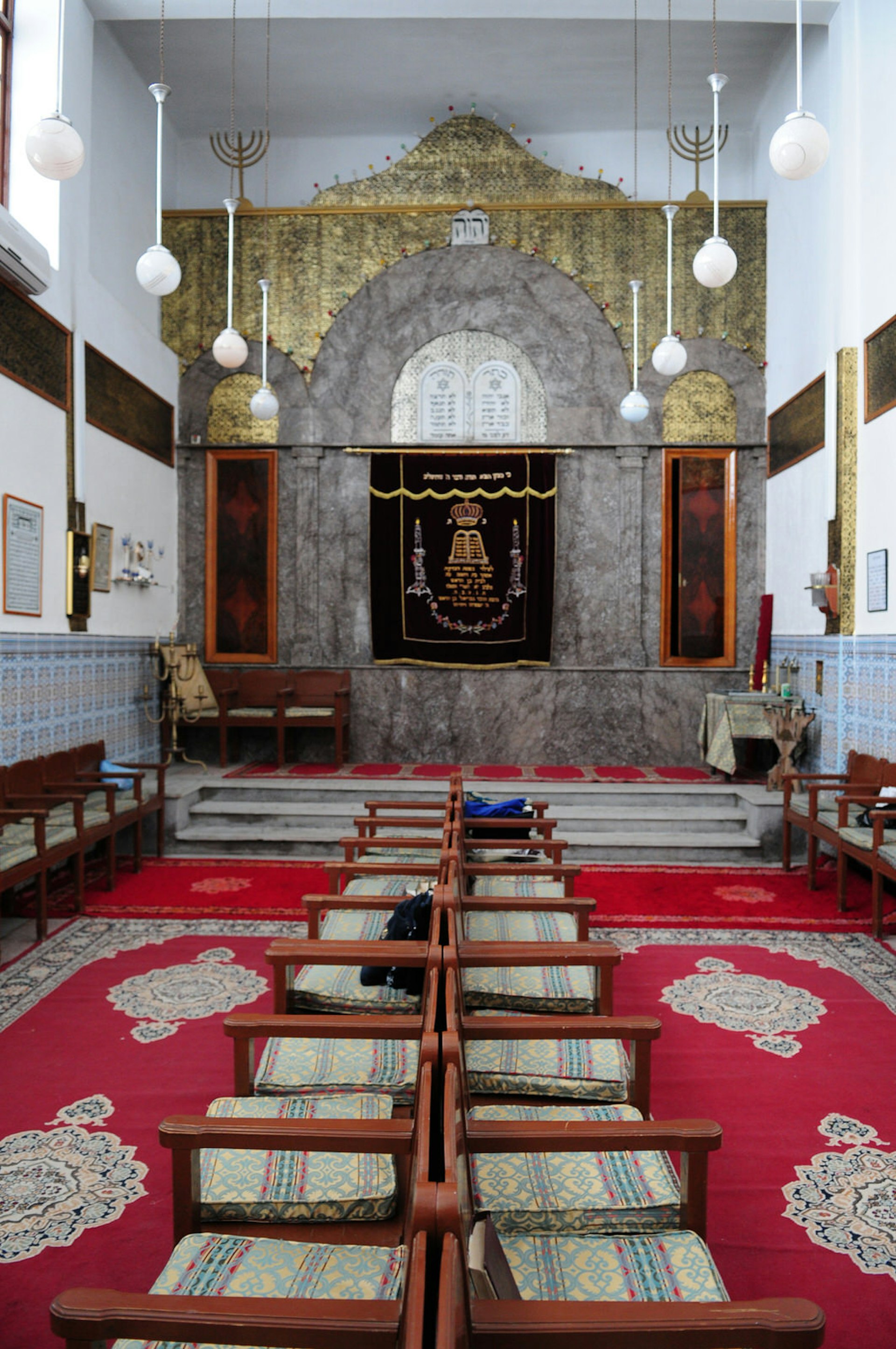 An inside view of the Lazama Synagogue, the oldest synagogue in the old Jewish quarter of Marrakesh, Morocco. Cushioned, wooden chairs sit on patterned red carpets. 