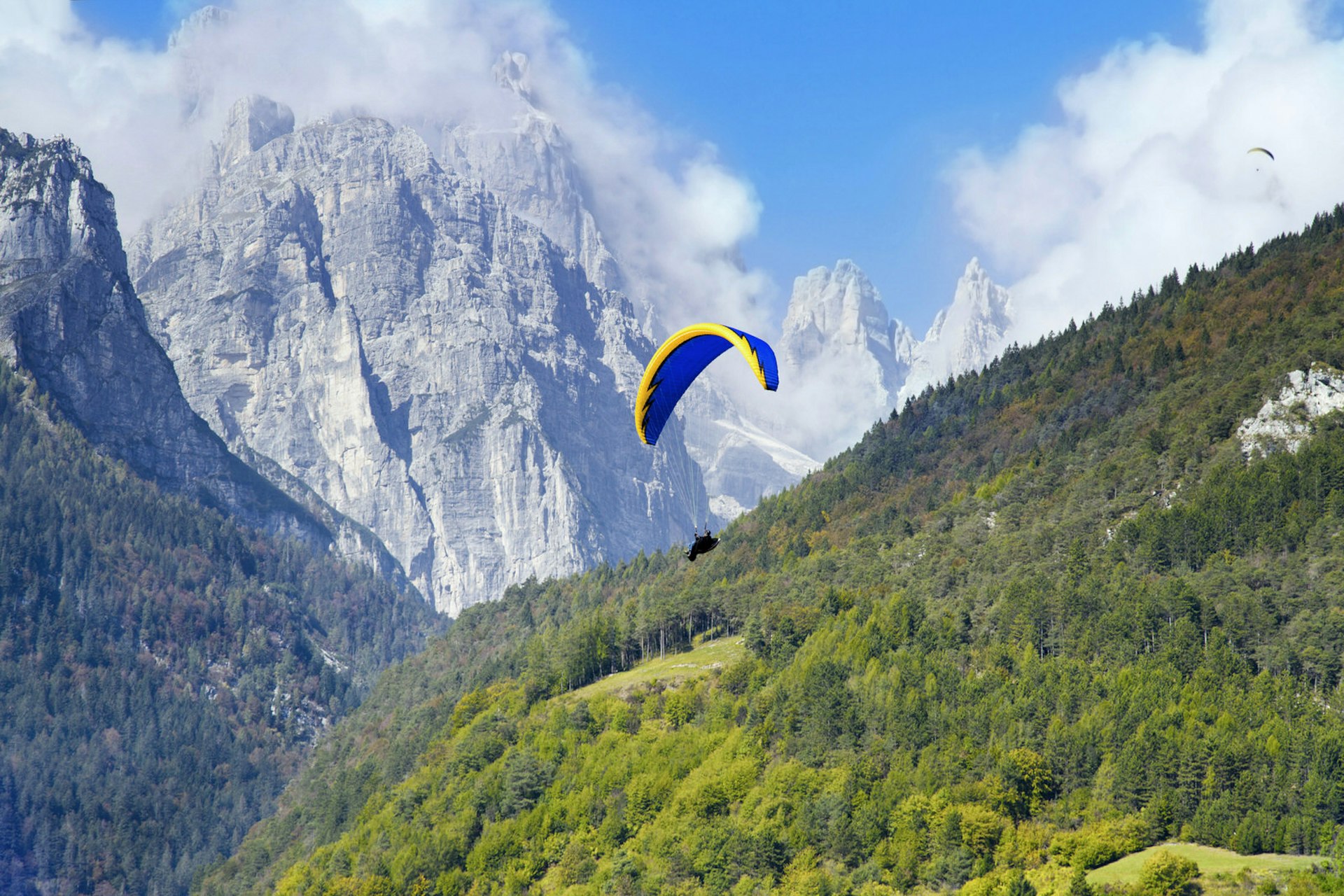 A paraglider flies past evergreen forests and rocky mountain peaks