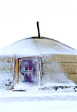 A yurt tent with a red door is covered in snow with a motorcycle iced over outside