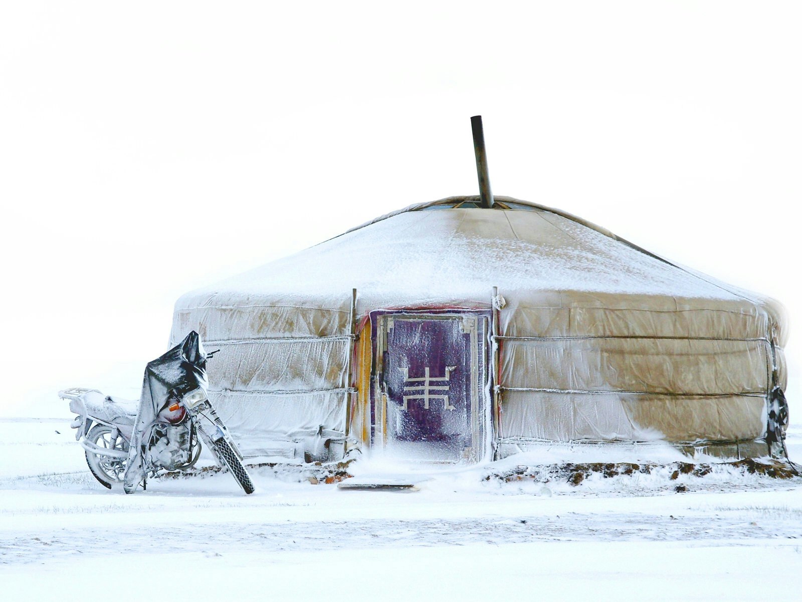 A yurt tent with a red door is covered in snow with a motorcycle iced over outside